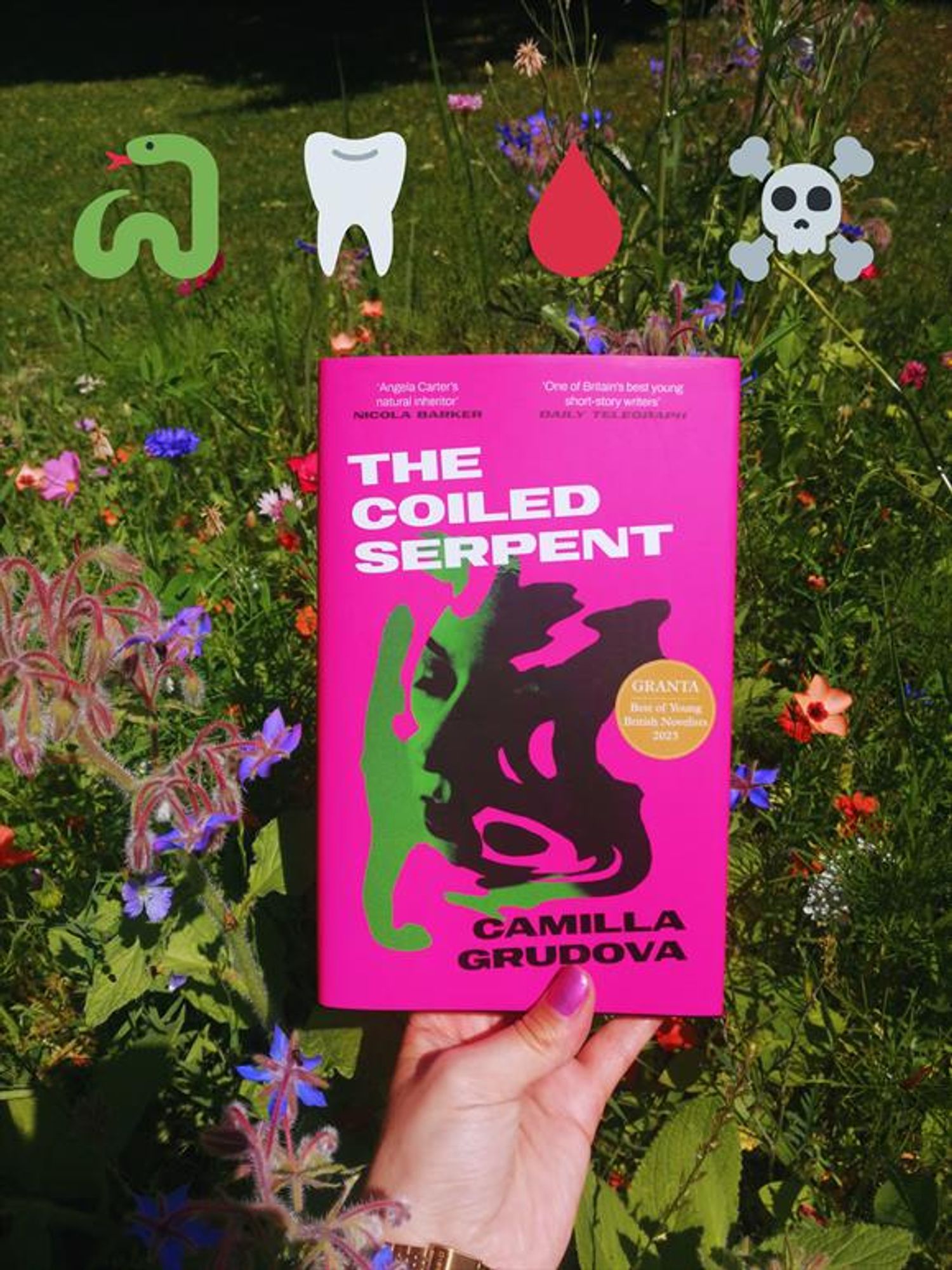 holding the short story collection "The Coiled Serpent" by Camilla Grudova in front of a wildflower meadow. 4 emoji decorate the image: a snake, a tooth, a drop of blood and a skull with crossed bones