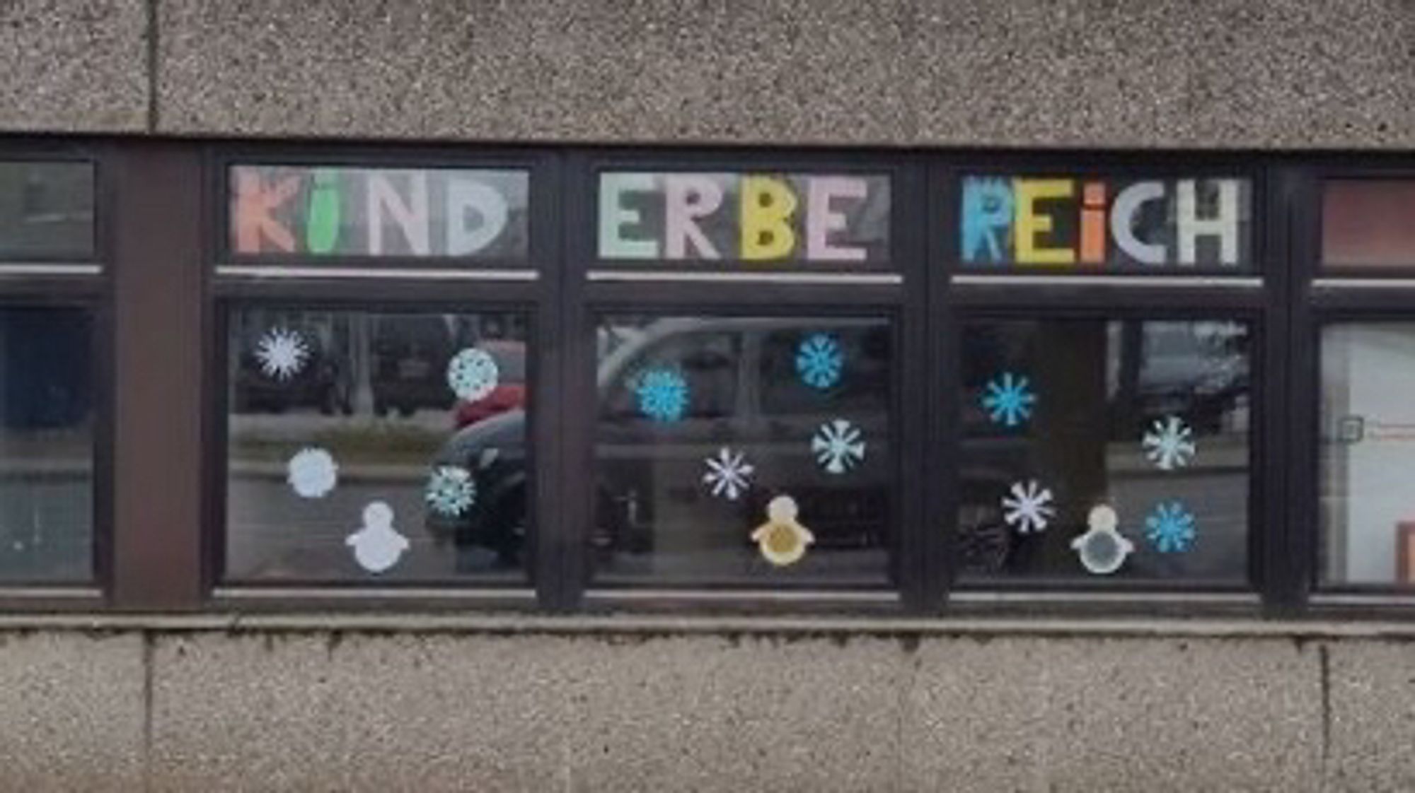 Drei Fenster in denen oben das Wort "Kinderbereich" in großen bunten Lettern prangt. Durch die drei Fenster liest es sich wie "Kind erbe reich".