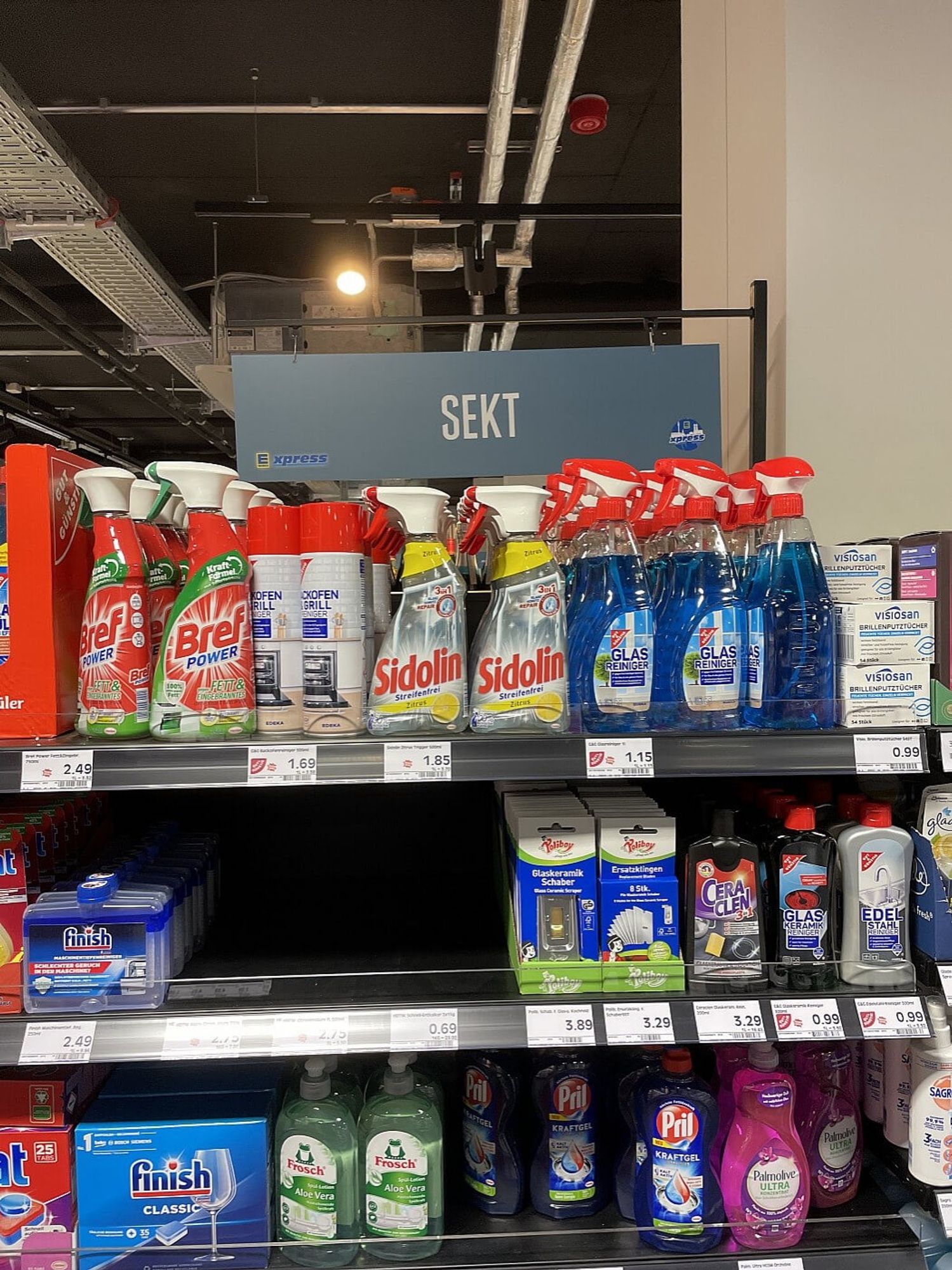 Ein Putzmittelregal im Supermarkt mit Glasreiniger, Spülmaschinenreiniger und Spülmittel. Darüber hängt das Schild "Sekt".