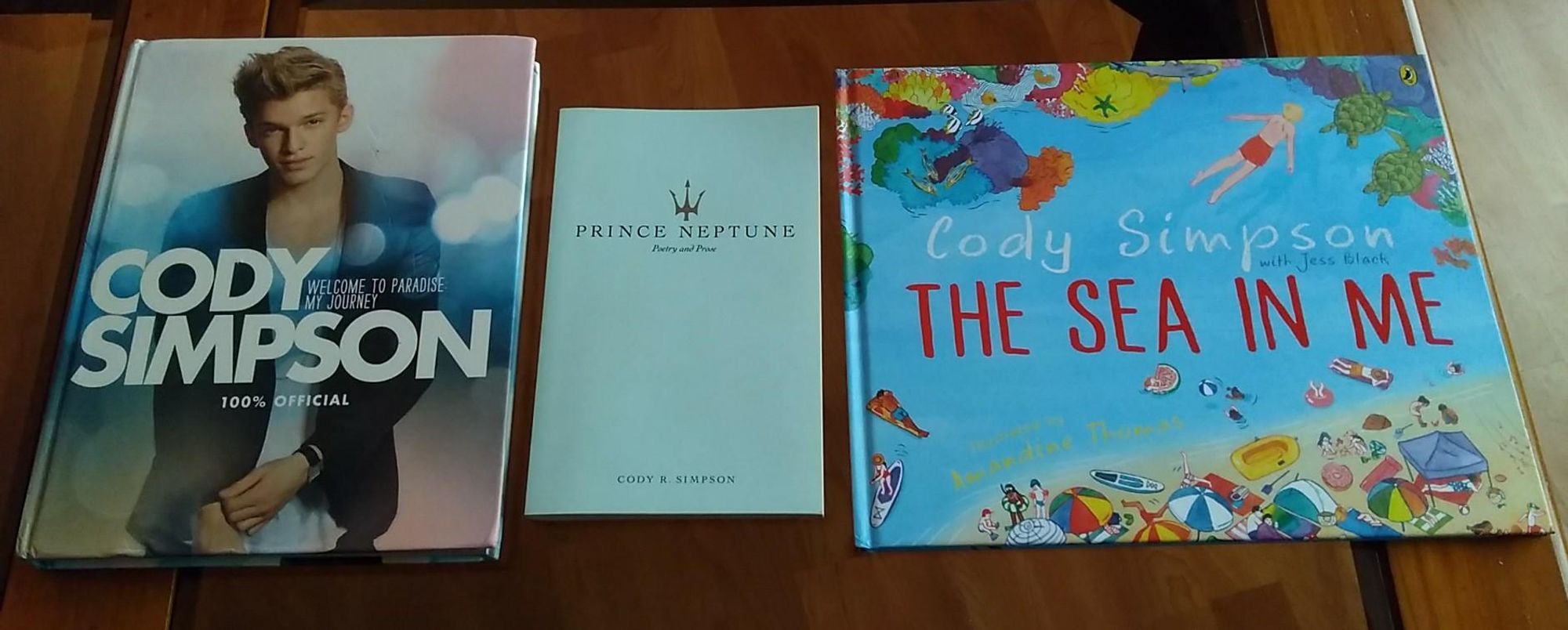 Colour photo of three books.
On the left is "Cody Simpson's Welcome To Paradise: My Journey" (ISBN 9780007520565), then "Prince Neptune: Poetry and Prose" (ISBN 9781524853990) in the centre, and "The Sea In Me" (ISBN 9781761049965) on the right. All three books are on a oak coffee table, with glass panel inserts.