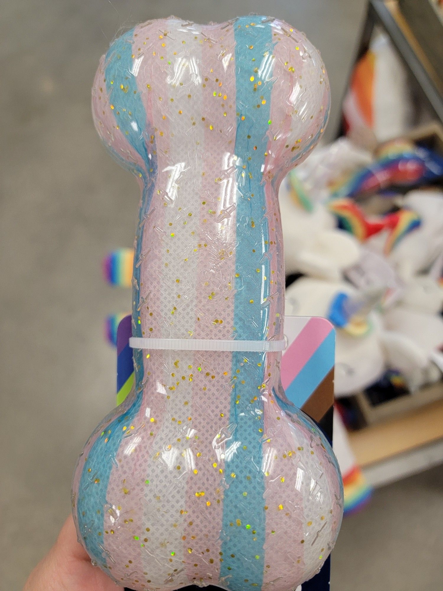 Pride themed dog toy at Petco, a semi-flexible transparent plastic cartoon bone with fabric visible within. The fabric has stripes of the Transgender pride flag (pastel pink, pastel blue, and white)