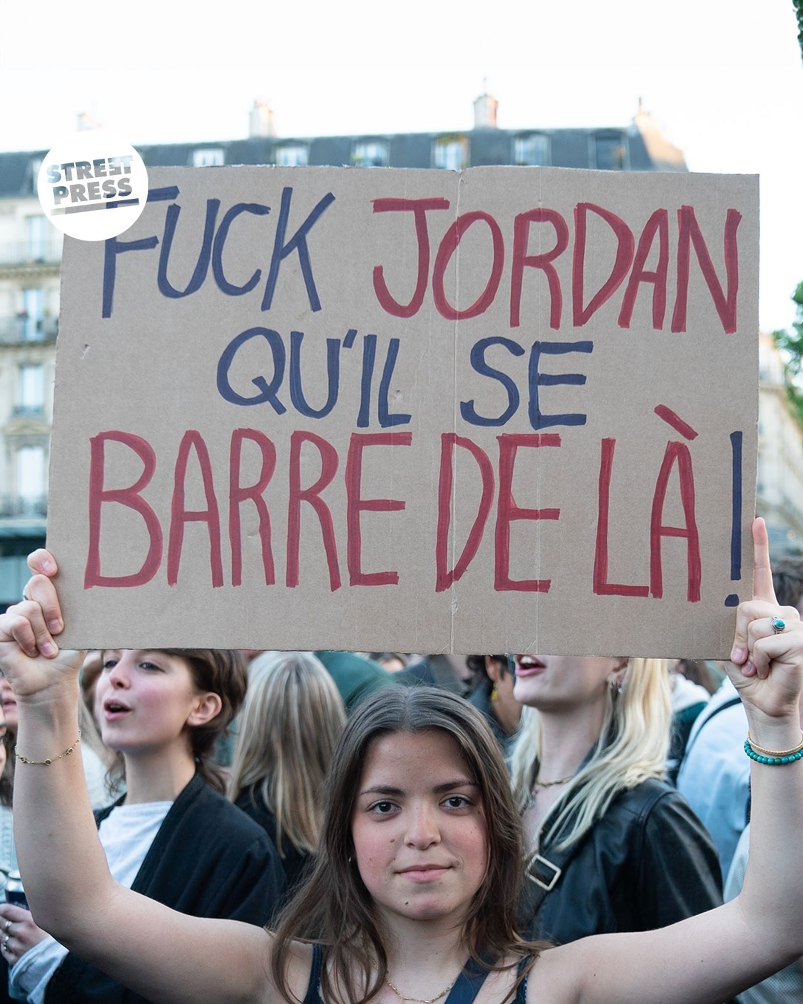 Jeune femme brune en manifestation qui tient une pancarte "Fuck Jordan qu'il se barre de là!" avec le logo StreetPress en haut à gauche