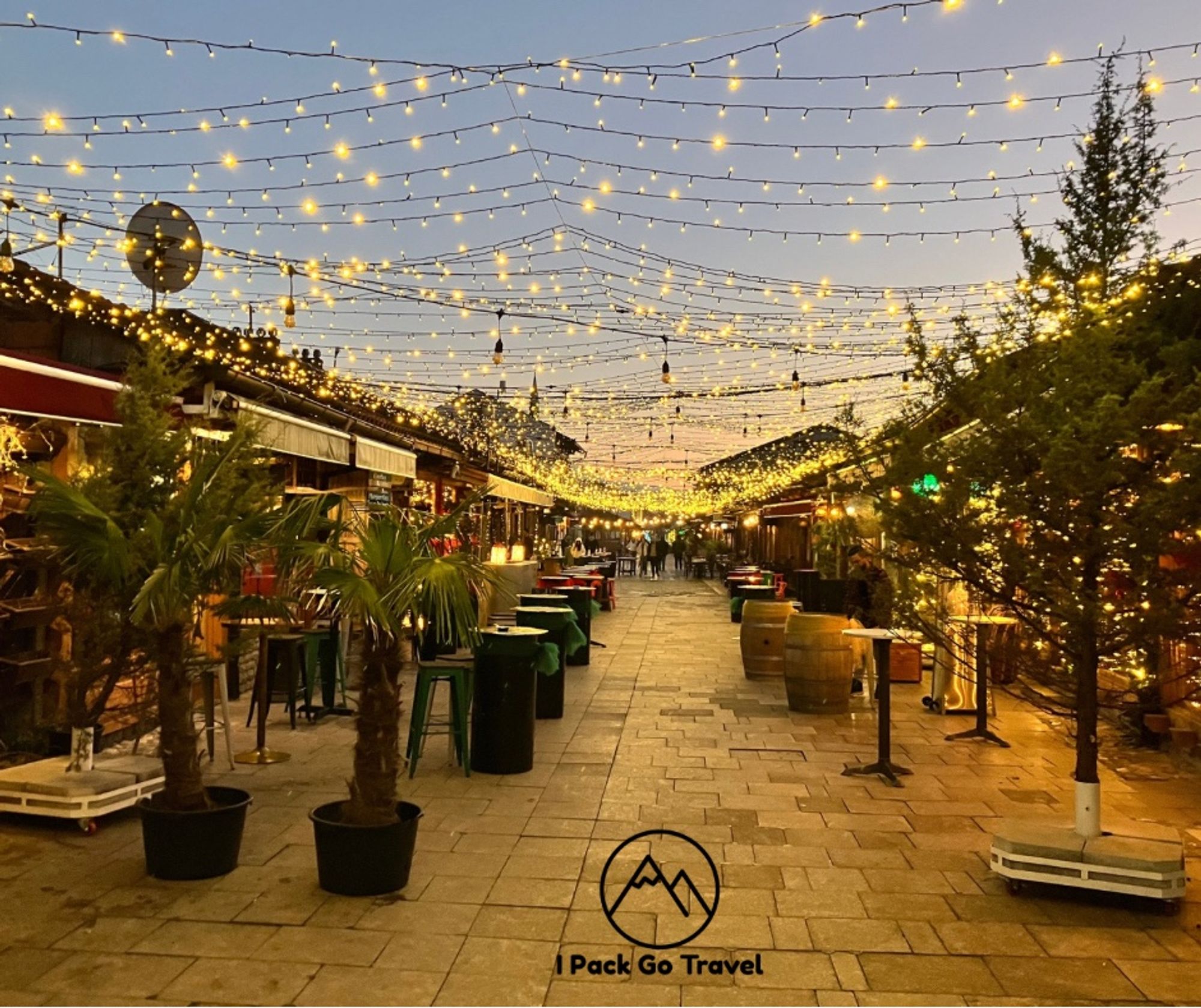 A festive lane of pubs, café bars with lots of outdoors seating and overhead fairy lights. There are trees and plants visible too. Pedestrians are vaguely visible in the distance. The evening twilight adds a cosy atmosphere to the image.