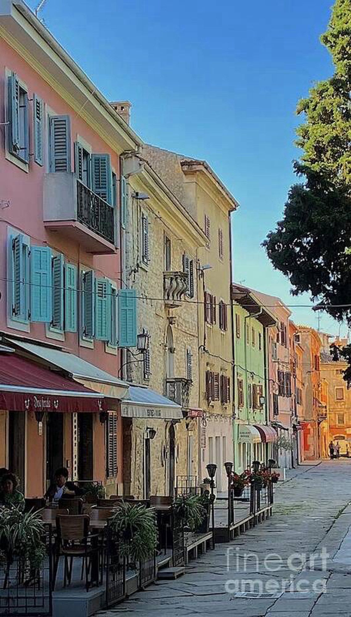 Street view of colourful buildings and window shutters. The street is paved with large bricks and there is blue sky when you look upwards. A street cafe with patio furniture, a canopy and vague silhoettes of patrons create a cosy atmosphere.