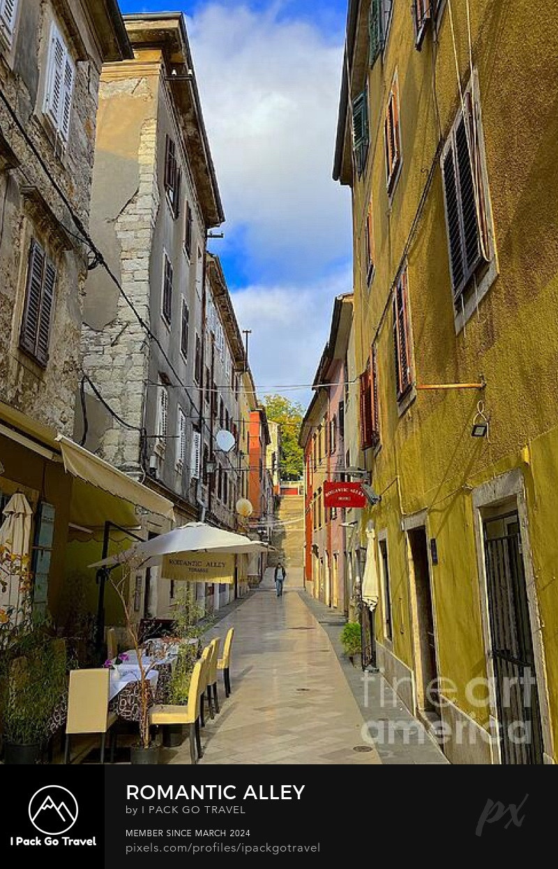 A romantic old town alley, with colourful weathered buildings, shutters, and a café with chairs, tables, patio umbrellas and a blue sky background. A pedestrian can be seen walking down the alley. The artist's logo and the theme title “Romantic Alley” are displayed in the footer.