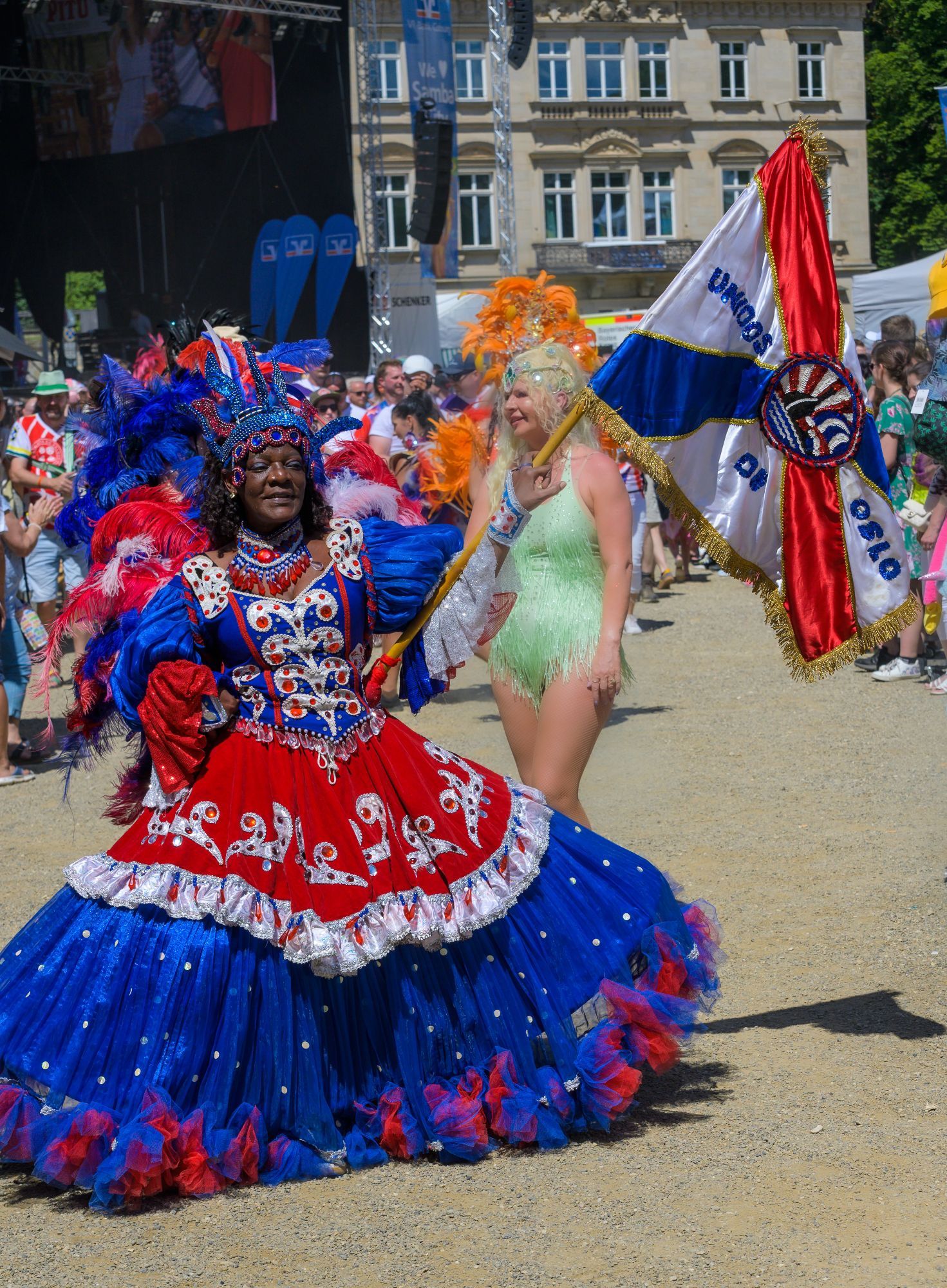 Auf dem Bild ist eine farbenfrohe Parade abgebildet. Im Vordergrund steht eine Frau in einem aufwändigen Kostüm, das in den Farben Blau, Rot und Weiß gehalten ist. Sie trägt ein weites Kleid mit Rüschen, Perlen und Glitzerelementen sowie einen beeindruckenden Kopfschmuck aus Federn in Blau und Rot. Ihre Pose zeigt Selbstbewusstsein und Eleganz.

Hinter ihr geht eine weitere Frau, die eine Fahne trägt, auf der "Unidos de Oslo" steht. Ihr Kostüm ist leichter und sie trägt ein ärmelloses grünes Kleid mit Fransen. Um sie herum sind weitere Menschen in Festkleidung, die an der Parade teilnehmen. Die Stimmung wirkt festlich und lebendig, es scheinen viele Menschen anwesend zu sein.

Im Hintergrund sieht man ein großes Gebäude mit mehreren Fenstern sowie eine Bühne, was auf ein größeres Event oder eine Veranstaltung hinweist.