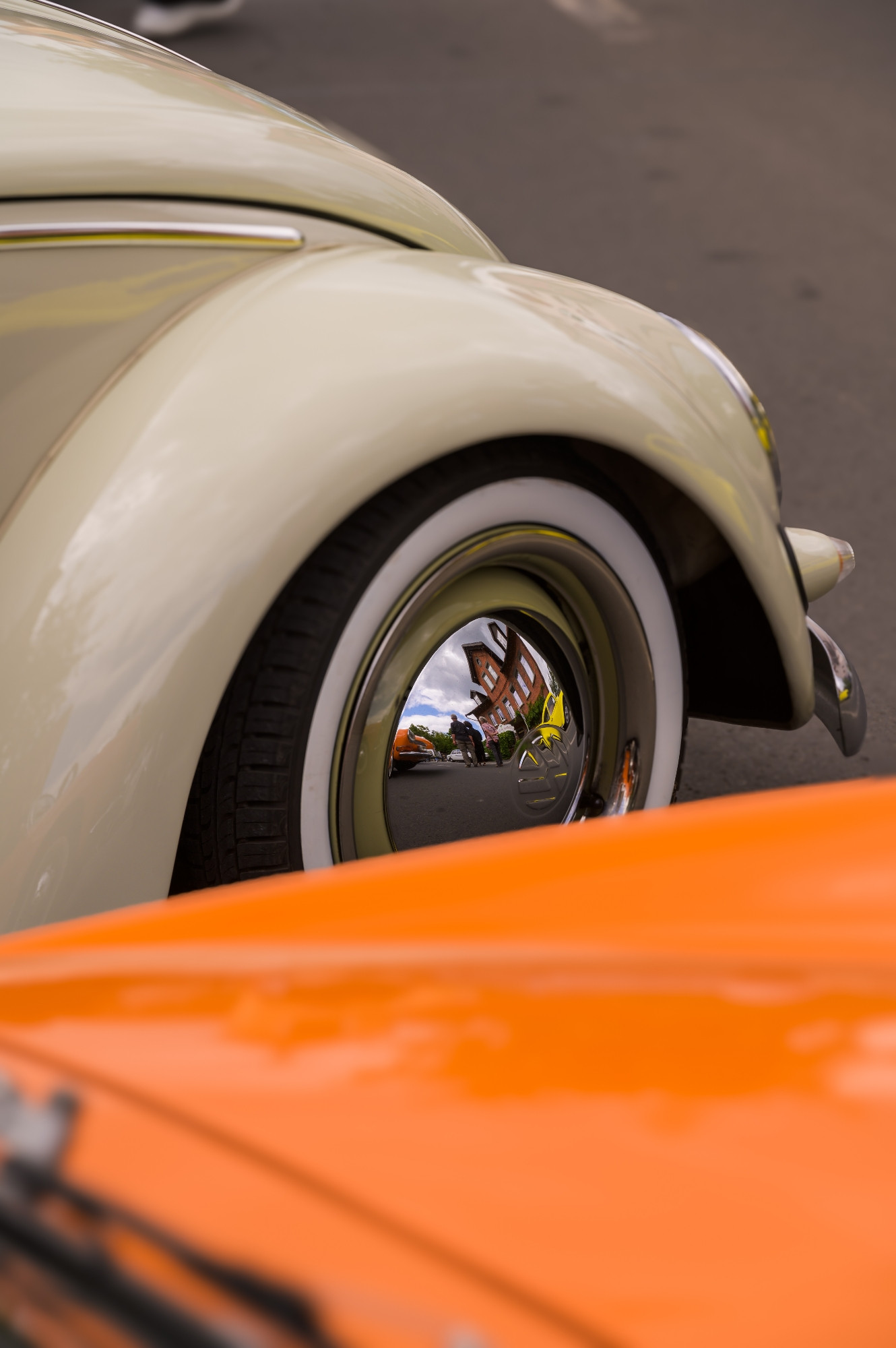 Das Bild zeigt einen cremefarbenen, klassischen Oldtimer mit einem besonderen Fokus auf das Rad und die Radkappe. In der glänzenden Radkappe spiegelt sich die Umgebung: Ein Gebäude, der Himmel und weitere Autos sind darin sichtbar. Im Vordergrund des Bildes ist ein orangefarbenes Fahrzeug zu sehen, das teilweise den unteren Bereich des Bildes bedeckt. Die Szene scheint auf einer Straße oder einem Parkplatz aufgenommen worden zu sein, möglicherweise bei einem Treffen für Oldtimer oder einer Autoshow. Die Details und die Reflexionen auf der Radkappe geben dem Bild eine interessante Perspektive und Tiefe.

