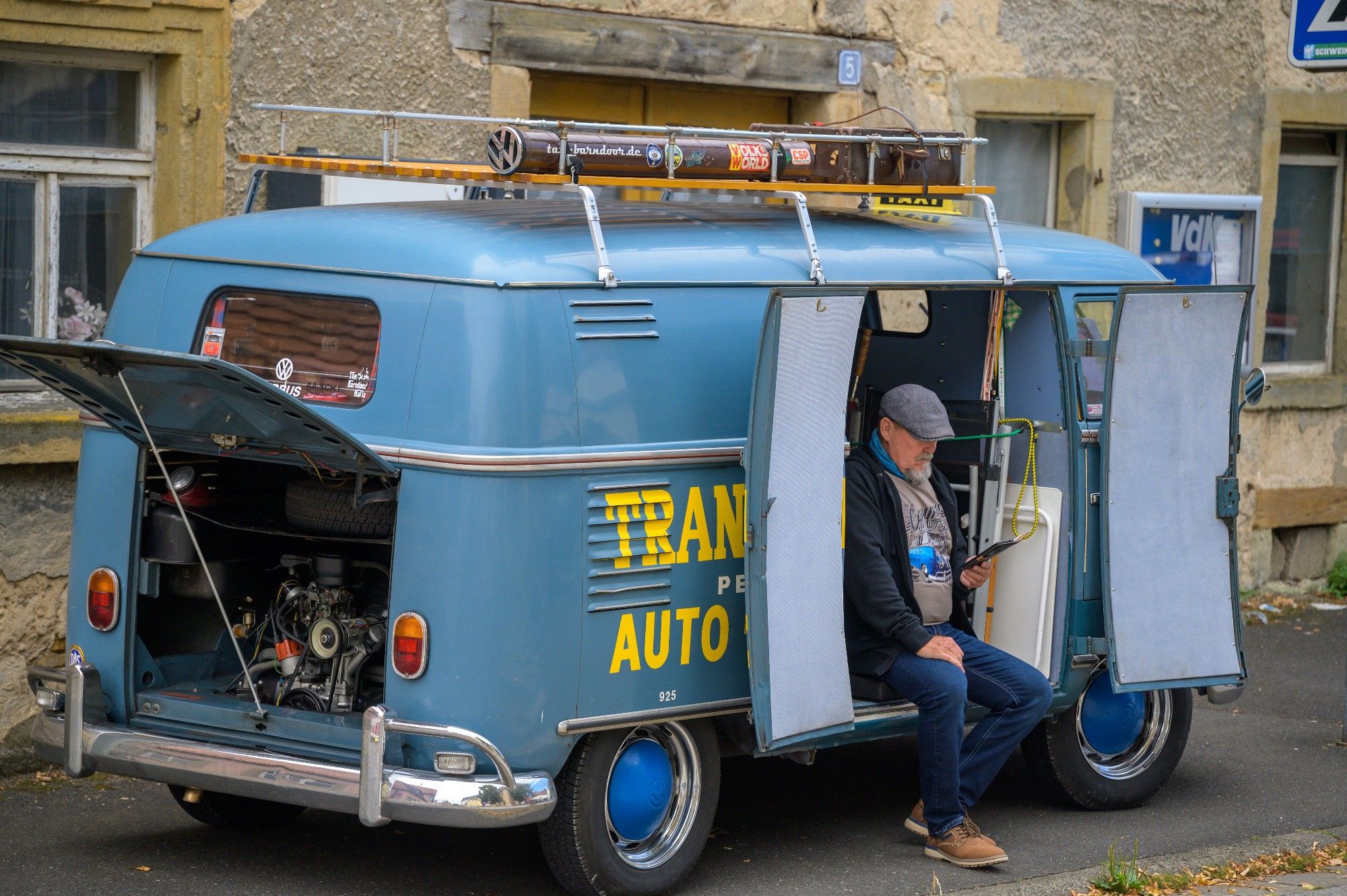 Auf dem Bild ist ein blauer, älterer Volkswagen-Transporter zu sehen, wahrscheinlich ein VW T1. Das Fahrzeug steht auf einer Straße vor einem Haus mit einer groben Steinfassade. Die Rücktür des Transporters ist geöffnet und der Motorraum ist gut sichtbar. Auf dem Dach befindet sich eine Gepäckablage mit Werkzeugen und Schildern.

An der Seite des Transporters steht in gelben Buchstaben die Aufschrift "TRANS AUTO", was darauf hinweist, dass das Fahrzeug möglicherweise zu einer Autoreparaturfirma gehört. Ein Mann mit grauem Haar und Bart sitzt auf der seitlichen Türöffnung des Transporters. Er trägt eine dunkelgraue Mütze, eine Jacke und eine blaue Jeans. In der Hand hält er ein Tablet oder ein anderes elektronisches Gerät, in das er vertieft zu sein scheint.

Die Umgebung wirkt ruhig, und es scheint sich um eine alte, ländliche Gegend zu handeln.