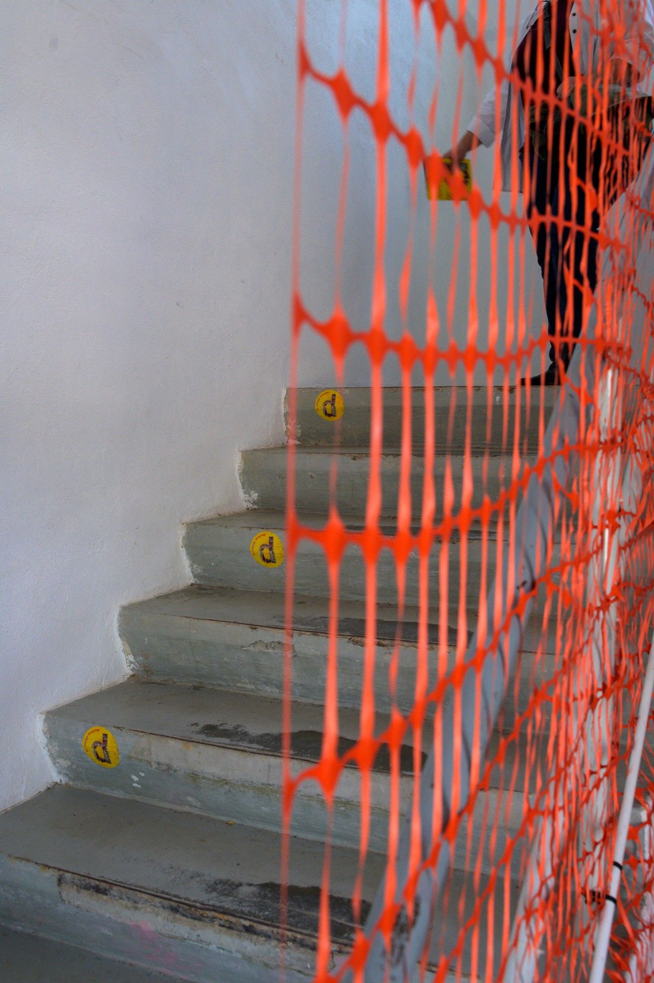 Das Bild zeigt eine Treppe, die von der Seite betrachtet wird. Ein leuchtend orangefarbenes Sicherheitsnetz verdeckt teilweise die Sicht auf die Treppe. Die Stufen scheinen aus Beton zu bestehen und weisen Gebrauchsspuren sowie einige Markierungen oder Flecken auf. An der Kante jeder Stufe sind gelbe Markierungen zu sehen, die möglicherweise als Maßangaben oder Warnhinweise dienen. Das Sicherheitsnetz ist an der Wand und dem Geländer befestigt, was darauf hindeutet, dass Bau- oder Reparaturarbeiten im Gange sind. Der Kontrast zwischen dem leuchtenden Orange des Netzes und den gedämpften Tönen der Treppe und der Wand hebt die Sicherheitsvorkehrungen in einer alltäglichen Umgebung hervor.