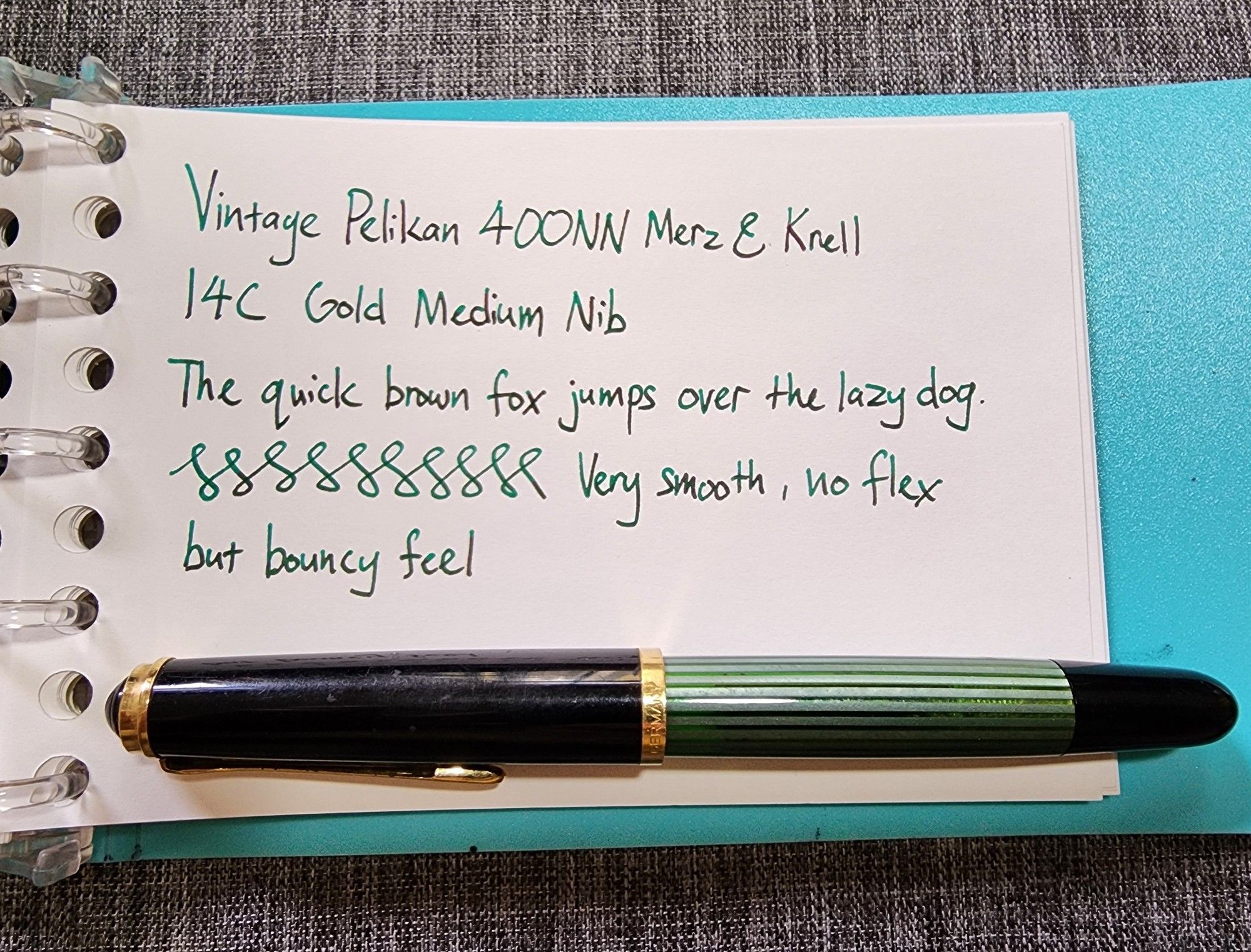 A green-and-black striped Pelikan fountain pen on a notebook page. There is handwritten text and some figure-eights drawn on the page with green ink. The text reads:
Vintage Pelikan 400NN Merz & Krell
14C Gold Medium Nib
The quick brown fox jumps over the lazy dog.
Very smooth, no flex but bouncy feel