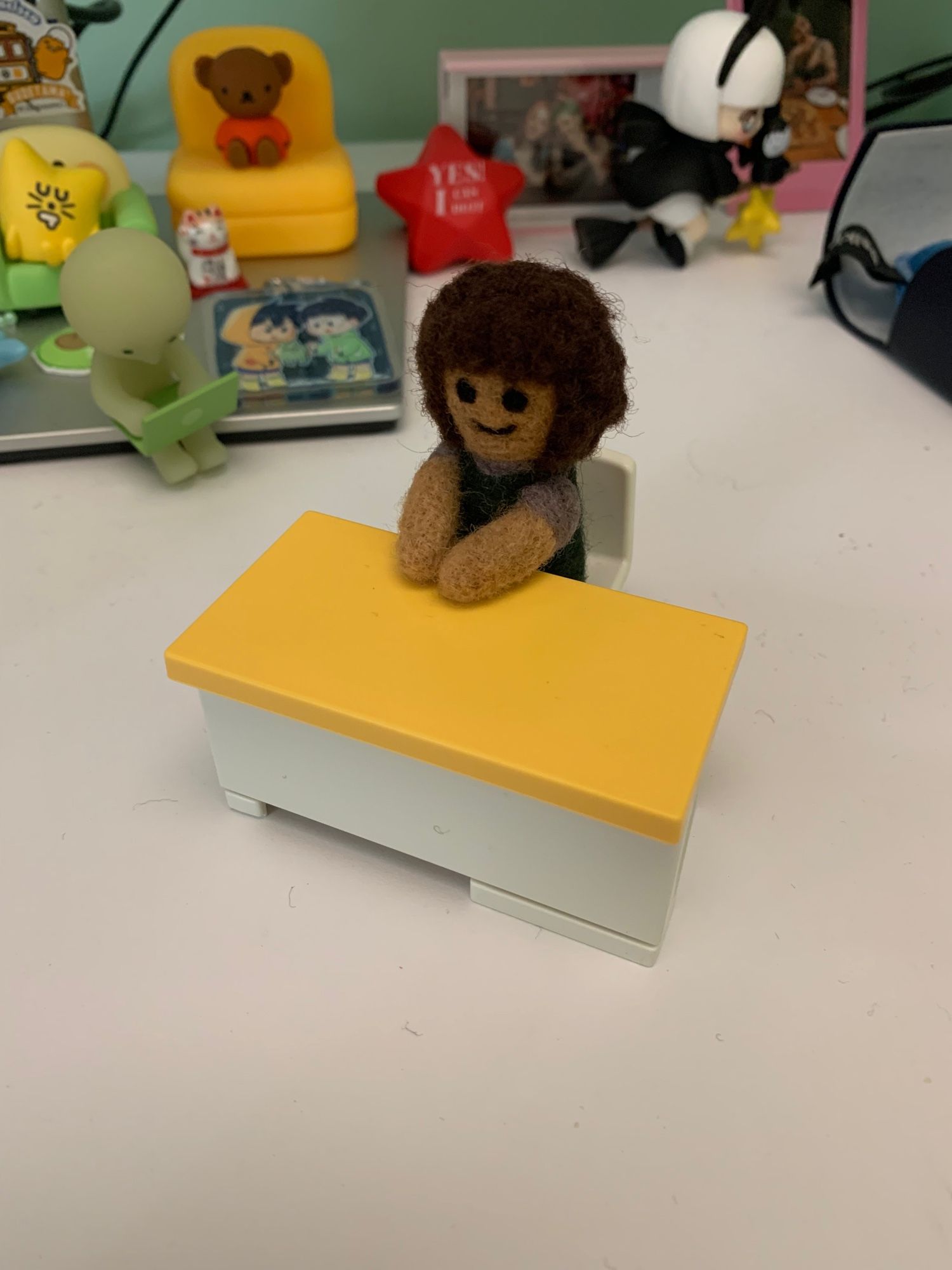 The same yellow and white toy desk but more close up. The white felted figure is replaced a curly haired brown girl in a green dress with grey sleeves. The implication is that the white figure has now been made into the girl.