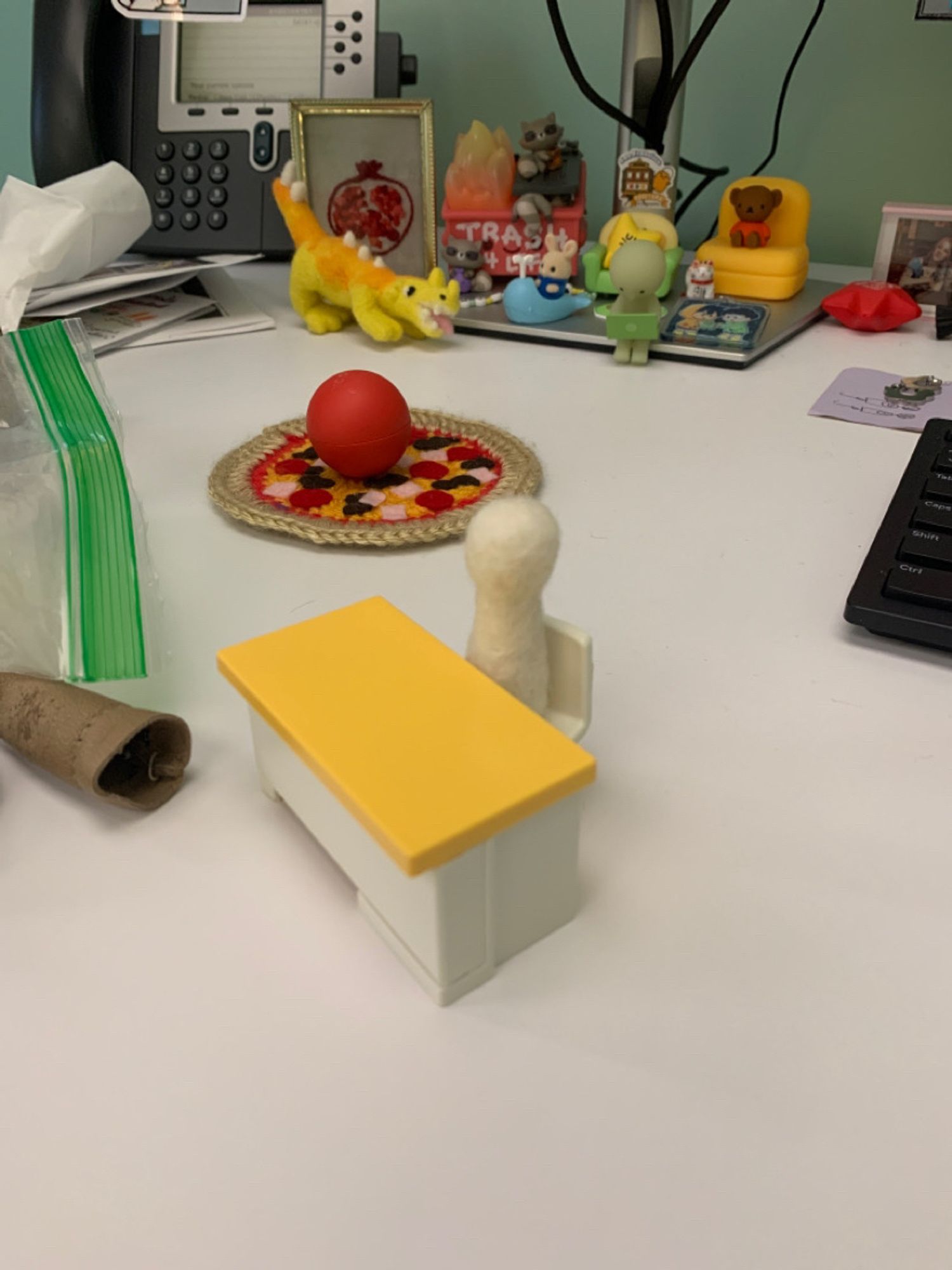 A small yellow and white toy desk with a vaguely human shaped white felt figure sitting at it. The toy is on top of a white desk with lots of clutter.