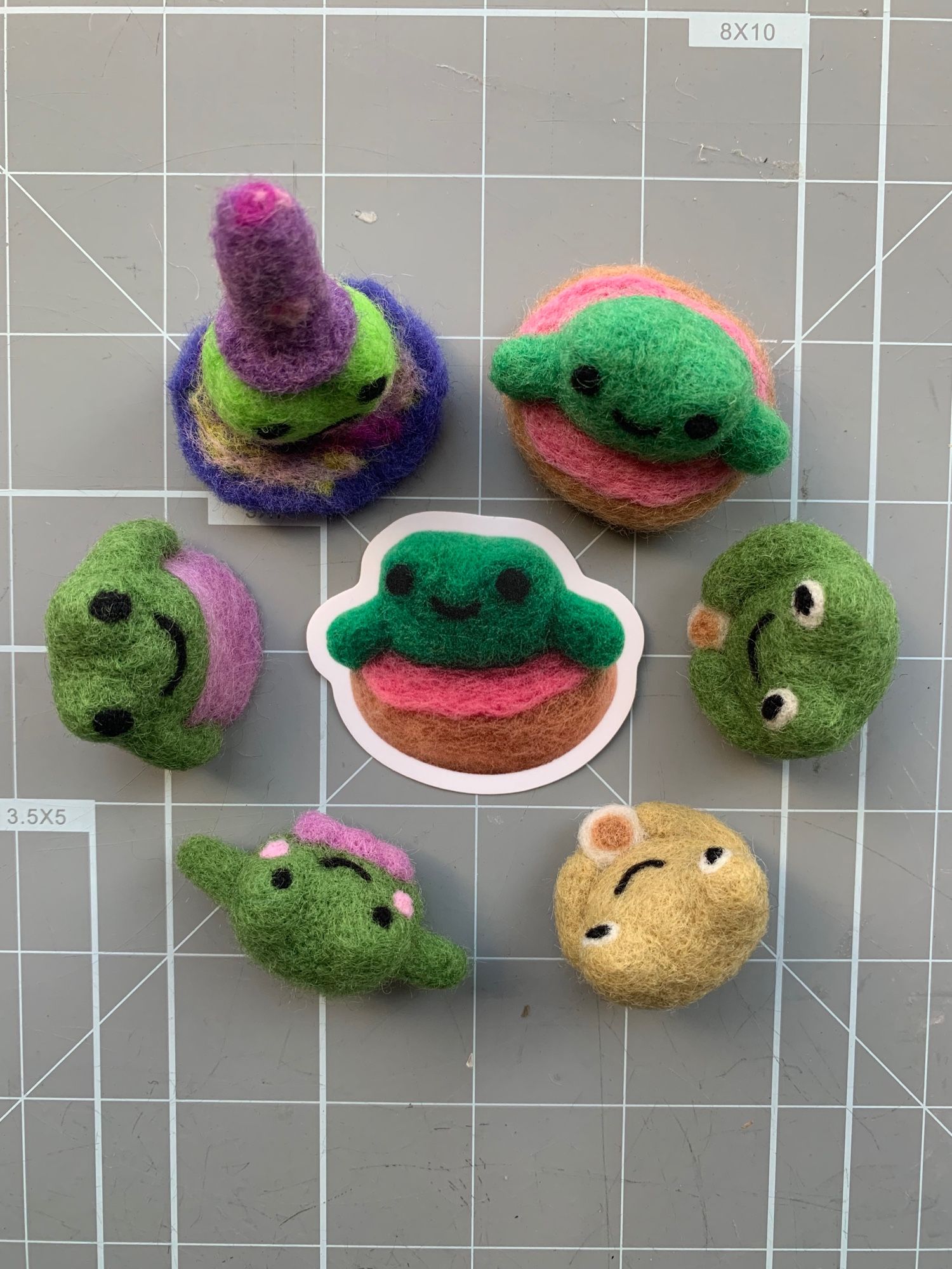 A circle of felted frogs (a wizard, frog in pink frosting donut floaty, two frogs with coffee, frog in purple bow tie, frog in purple dress) looking in at a sticker of the frog in a floaty