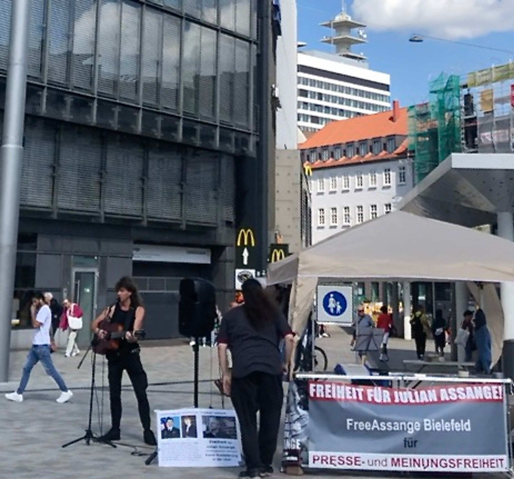 Ein Mann mit Gitarre steht vor einem Mikro, daneben ein Infostand mit dem Banner: "FREIHEIT FÜR JULIAN ASSANGE! FreeAssange Bielefeld für PRESSE- und MEINUNGSFREIHEIT"