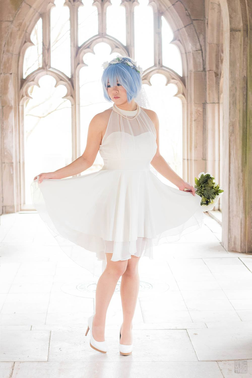 A woman with short light blue hair with a flower hairband in a white dress, veil and white heels holding a white bouquet and posing her dress in an outdoor church setting. She is cosplaying a wedding dress version of Rem from Re:Zero