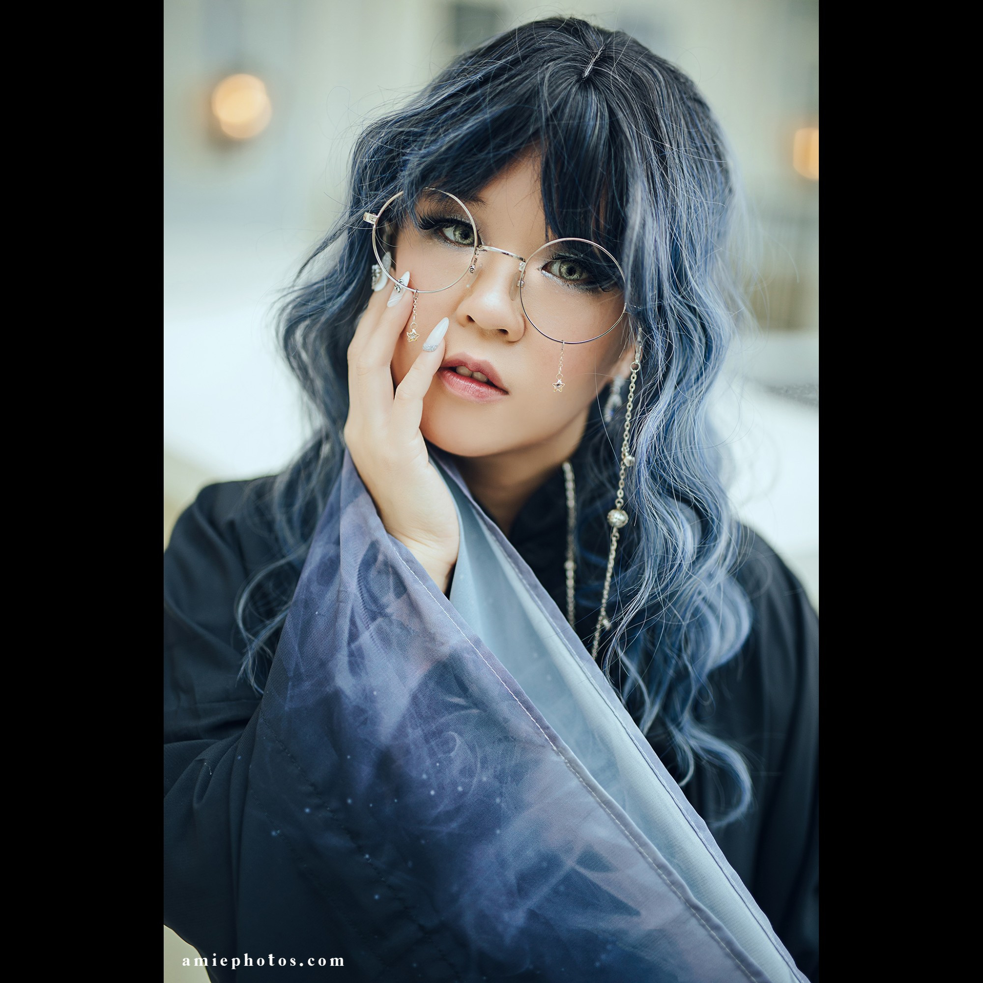 Photo of a woman with black blue gray hair with fashion glasses and bright grey eyes staring out with her right hand holding her face.  Her hand has long white and silver nails.