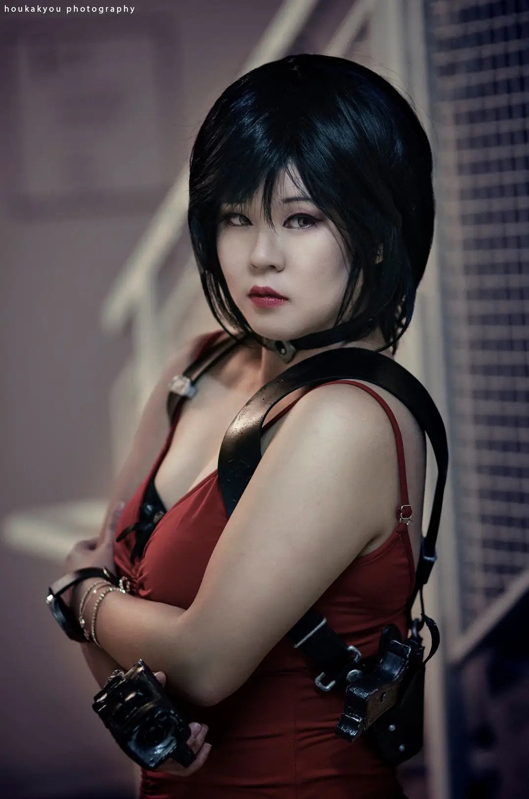 Asian woman cosplaying as Ada Wong from Resident Evil 2 Remake. She is wearing a red spaghetti strap dress and arms crossed, wearing a prop gun holster and holding a prop gun. She has short black hair, light brown eyes with dark lips and staring straight ahead.