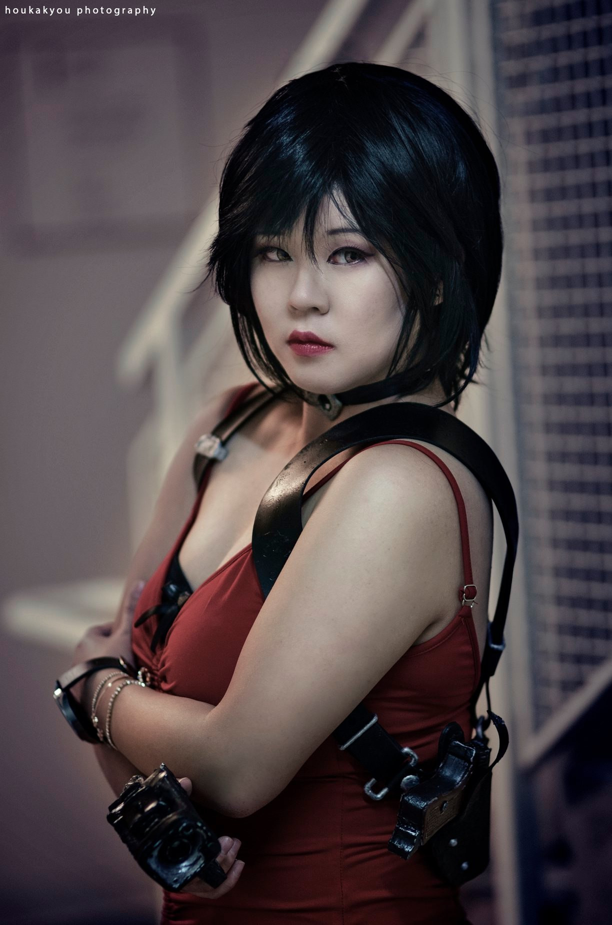 A woman cosplaying Ada Wong from the Resident Evil 2 remake. She is looking at the screen and has short black hair wearing a spaghetti strap red dress with a prop gun holster and holding a prop gun. 