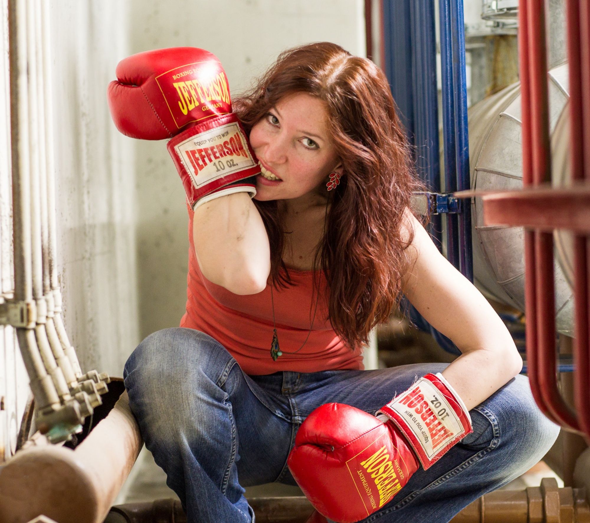 Fee kauert in der Hocke und zieht sich Boxhandschuhe an. Sie fletscht dabei die Zähne, um den Klettverschluss der Handschuhe mit den Zähnen enger zu ziehen. Ringsum ein industriell aussehender Innenraum mit blankem Beton, Rohren und Kabeln.