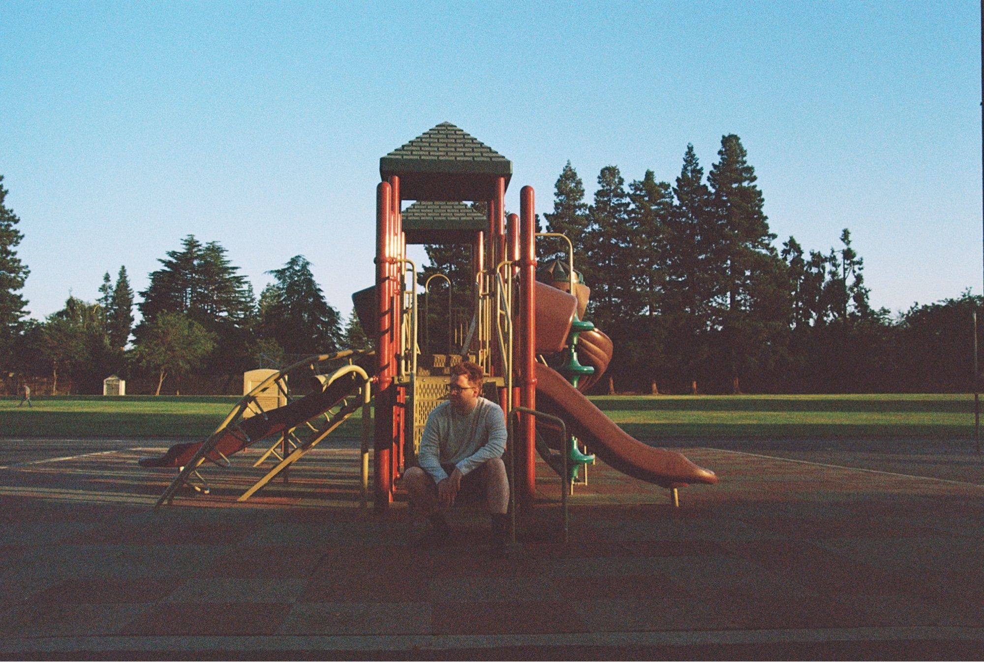 A man sits on the steps of a play structure. The sun illuminates the structure and the field behind and catches on the side of his face.