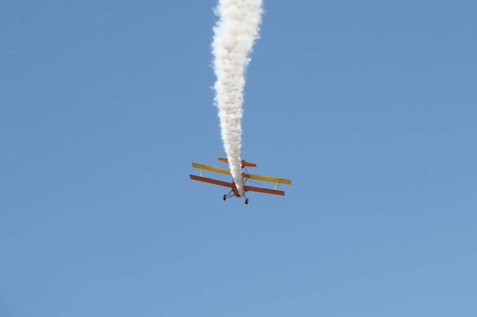 a skywriting biplane seen from below and behind against a blue sky with a plume of skywriting smoke coming from it