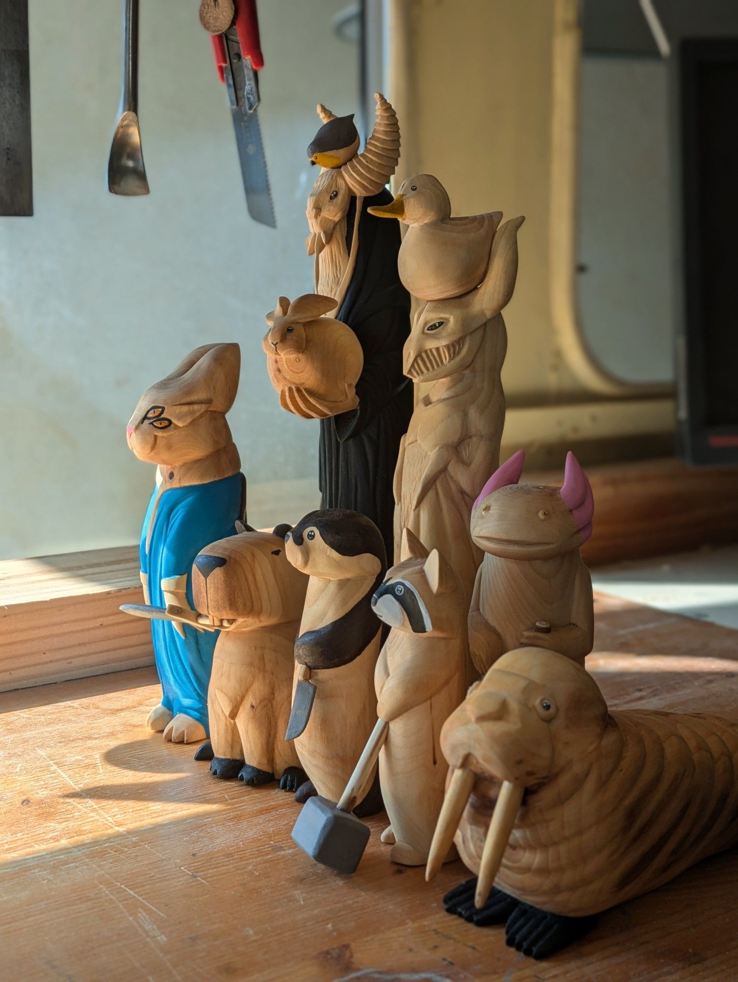 Woodcarved creatures on a workbench. A cat with horns, 4 eyes, goblins ears in a blue dress, a monster with 4 teeth and 2 horns with a rabbit on its left hand and a red robin on the top of its head, wearing a black dress, a dragon-like creature with a duck on the top of its head, a capybara wielding a sword between its teeth, an otter with a knife, a raccon with a sledgehammer, a big walrus and an axolotl holding an ax (wich is not visible on the picture).