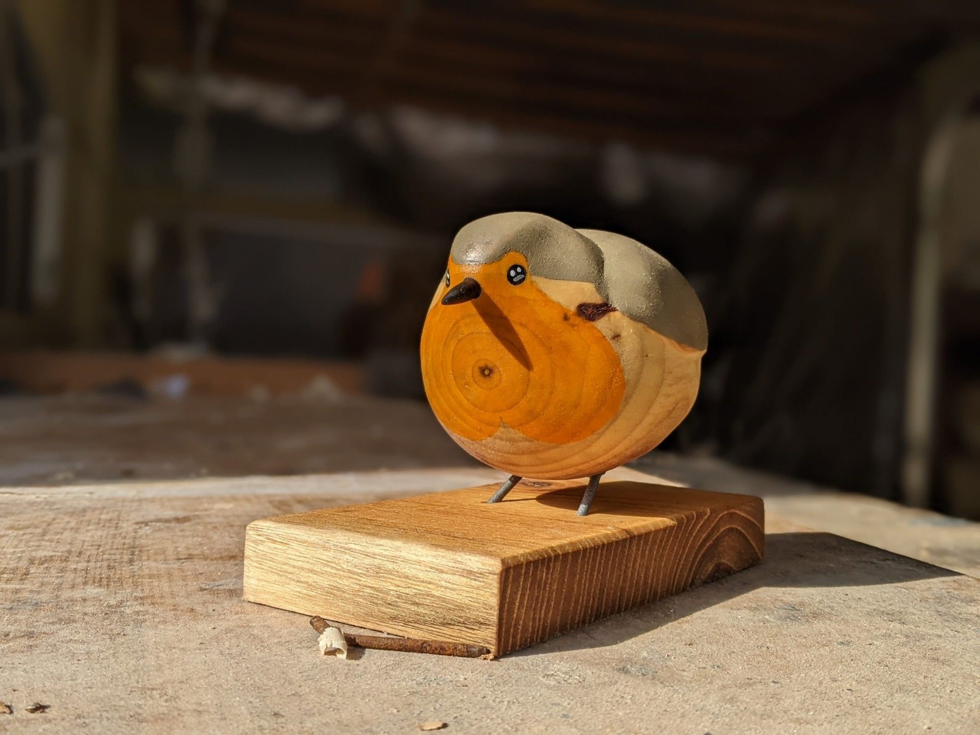 Sculpture sur bois d'un rouge gorge dodu, sur socle. Ses pattes ne sont pas détaillées se sont deux tiges en fer le rattachant au socle. Il est très rond. Il est peint en orange pour sa poitrine, son bec et noir, deux yeux tacheté de blanc et gris pour donner un éclat de vie, et du gris pour le haut de sa tête et son corps. Il est posé sur un plan de travail.