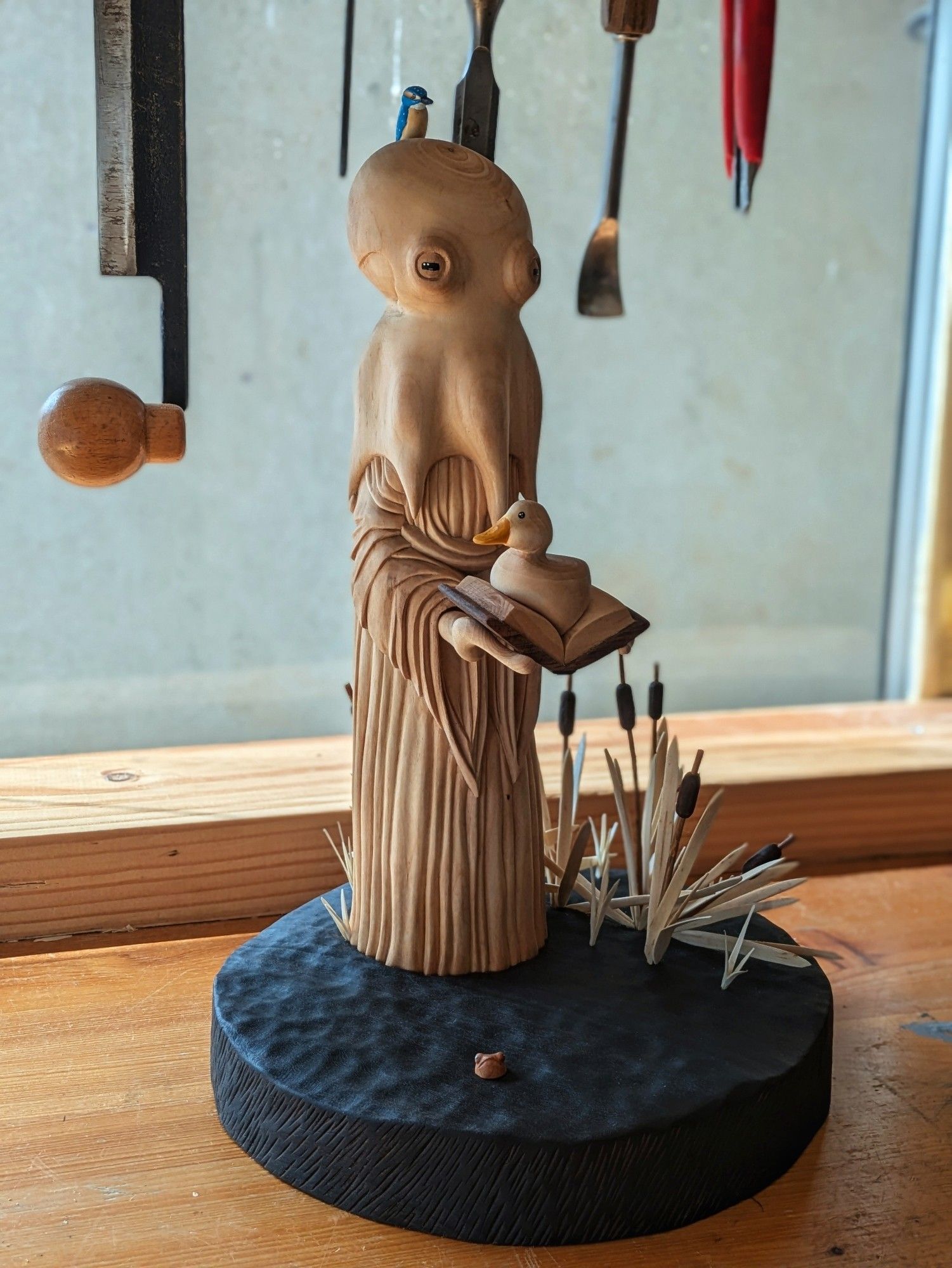 Woodcarved scene of a humanoid octopus wearing a dress on the surface of a pond. The creature has a kingfisher on its head. It's holding a book wich has a duck sitting on it. On the left of the base looking like water, bulrushes. On the right, a little frog has its head out of water. In the background woodcarving tools.