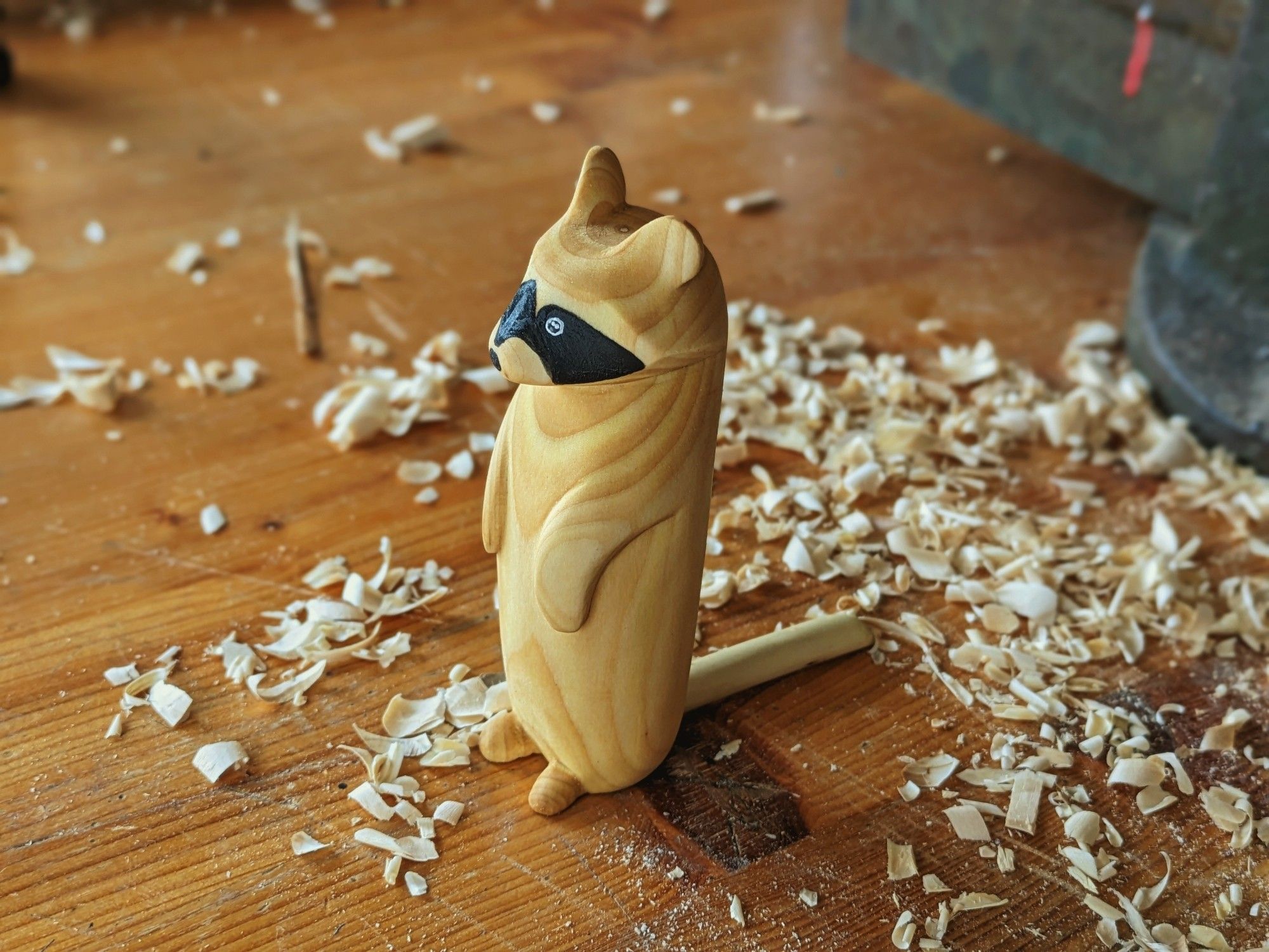 Woodcarved racoon on a workbench with wood chips all arround. It's standing up. The nose and the "mask" are paint in black