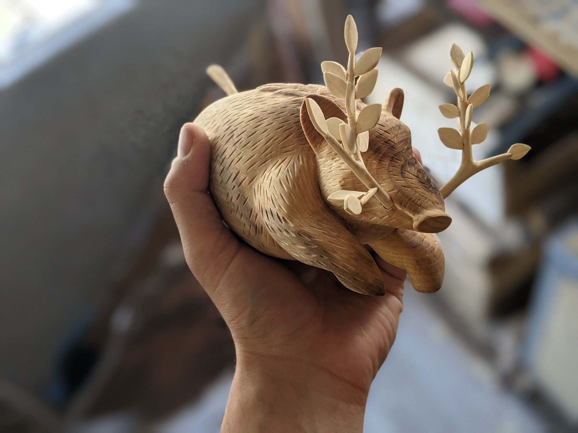 Sculpture sur bois d'un sanglier dont les défenses sont en fait des branches en feuilles. Il a une posture bondissante tout en étant rondelet. Des gravures suggèrent ses poils. Il est dans une main, vu de 3/4.