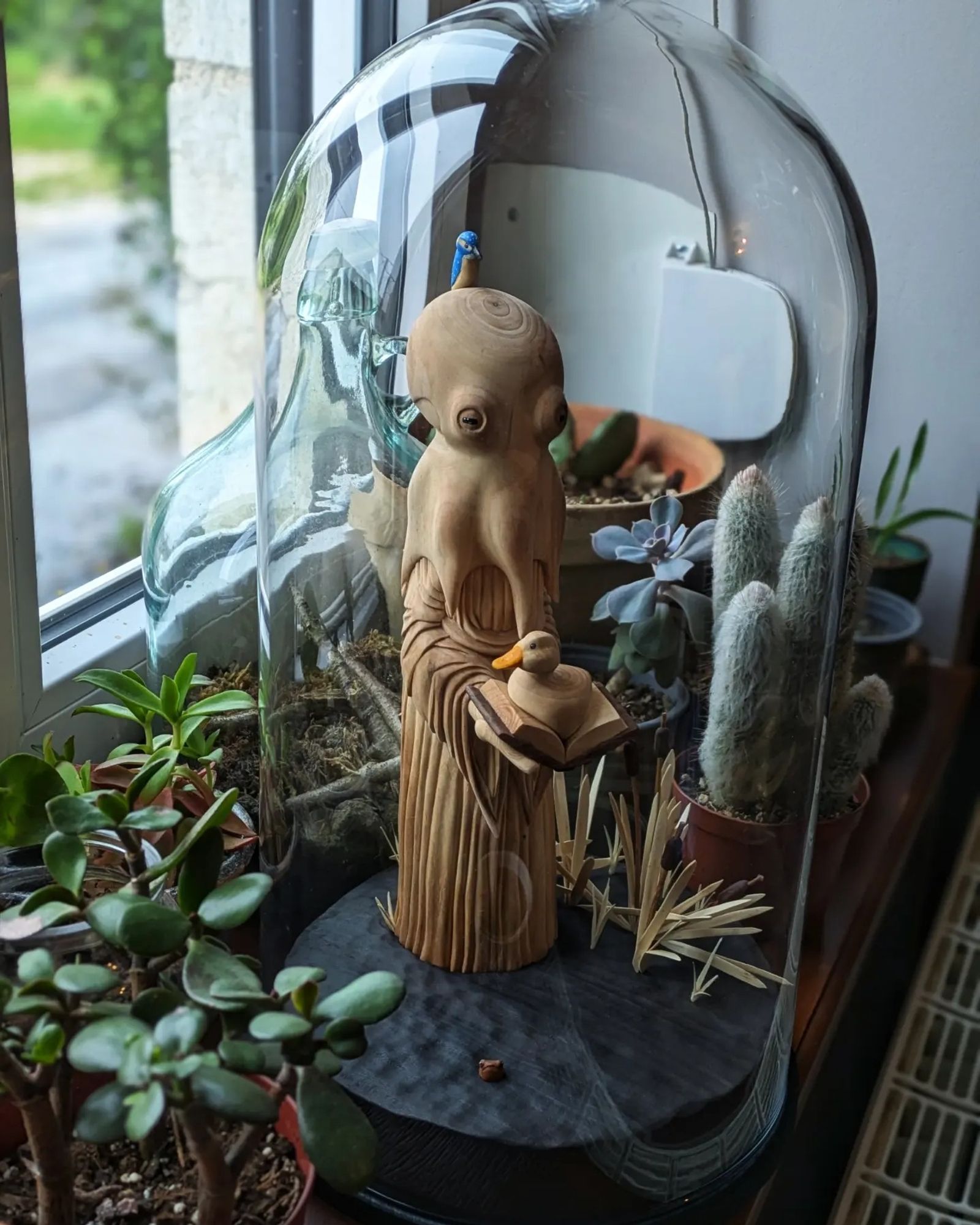 Glass bell with a woodcarved scene of a humanoid octopus wearing a dress on the surface of a pond. The creature has a kingfisher on its head. It's holding a book wich has a duck sitting on it. On the left of the base looking like water, bulrushes. On the right, a little frog has its head out of water. In the background, indoor plants by a window.