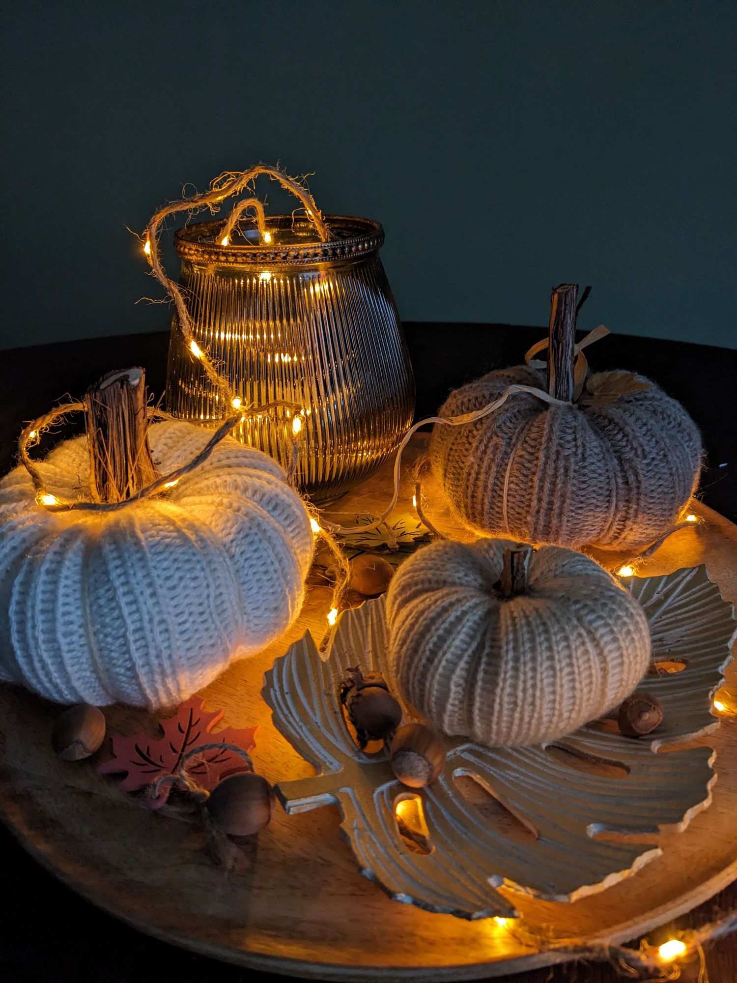 The picture shows knitted pumpkins on a plate with a wooden leaf and a string of lights.