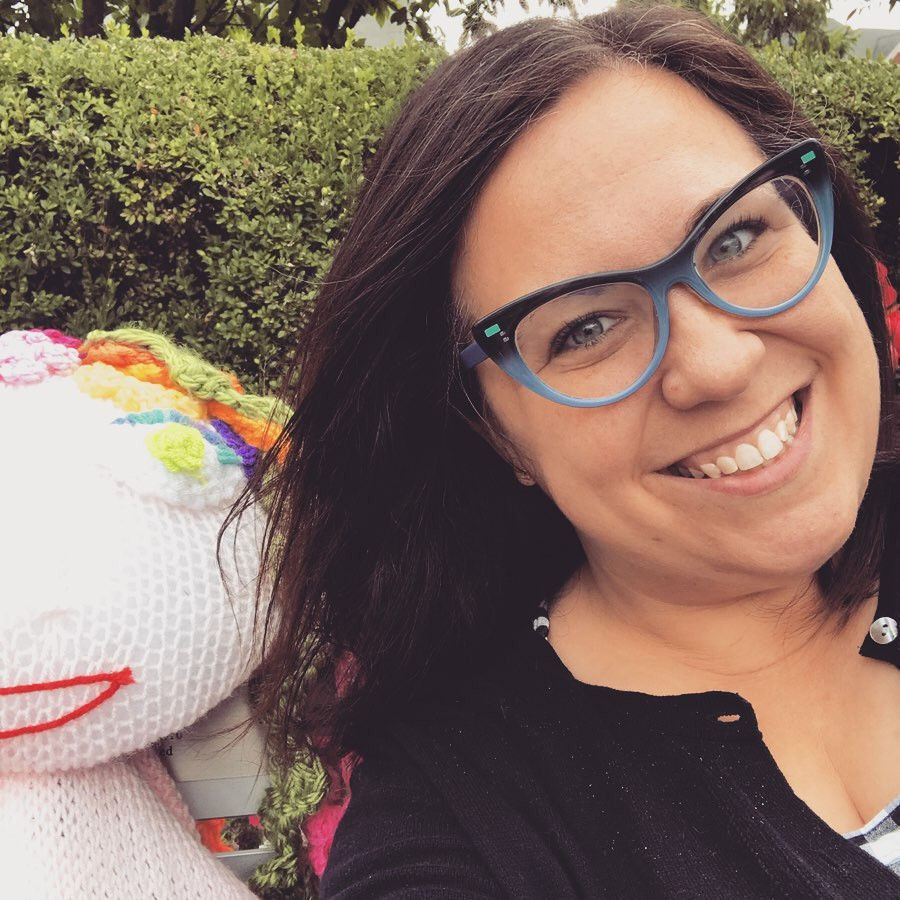 Woman in cat-eye glasses sitting next to a yarn-bomb art installation of a woman with rainbow hair.