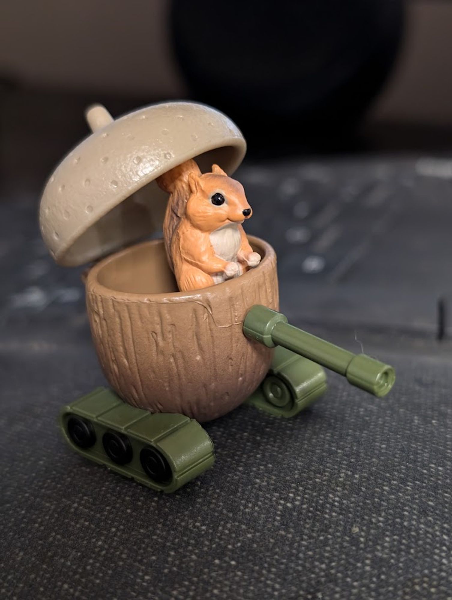 A toy figure of a small squirrel sitting inside a round, brown, wood-textured vehicle shaped like a half acorn. The acorn has a hinged top, partially open, and the vehicle is mounted on green tank treads with a small green cannon extending from the front. The background appears to be a soft, dark surface.