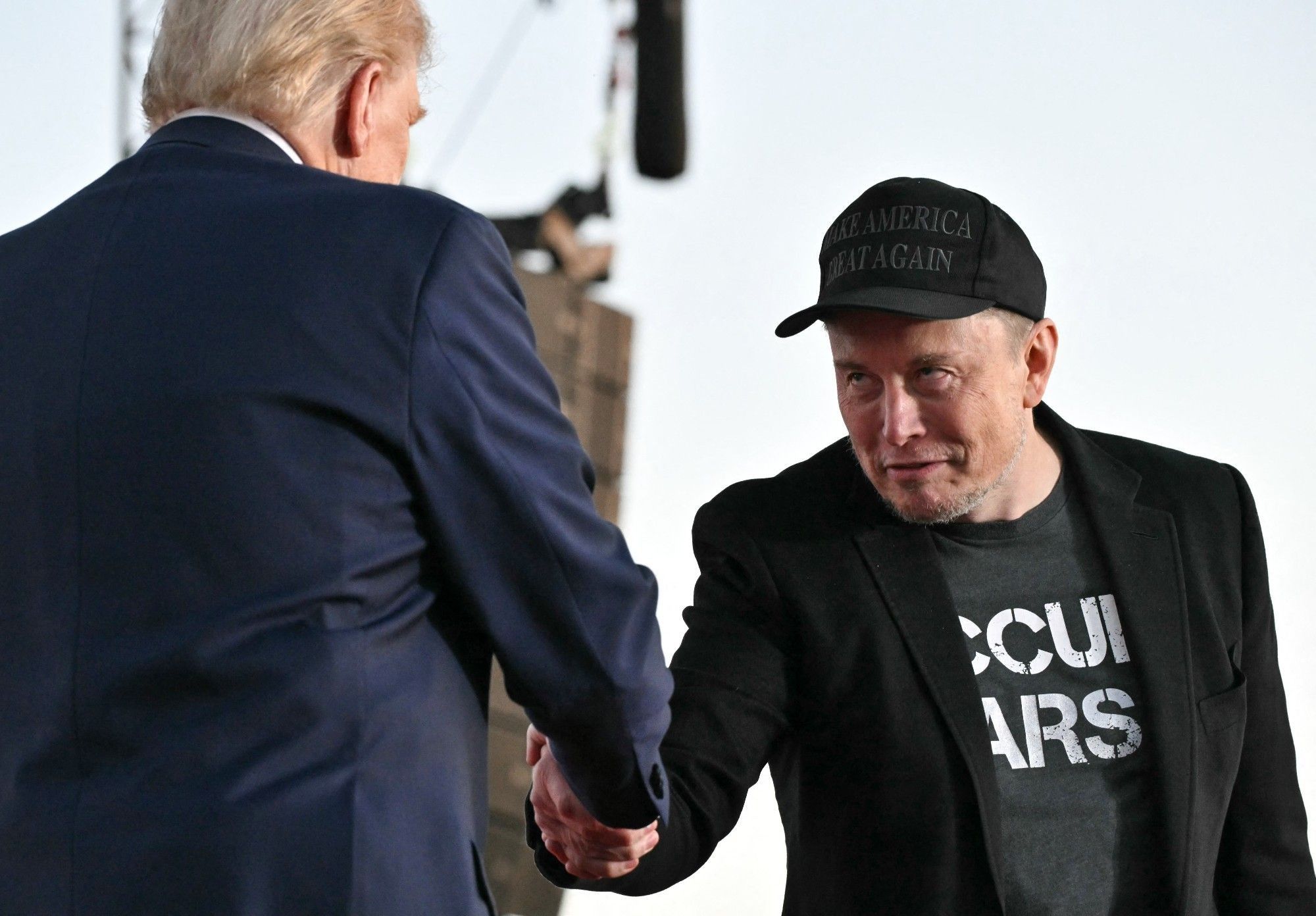 Musk shakes Trump's face. He is wearing creepy Psycho smile.