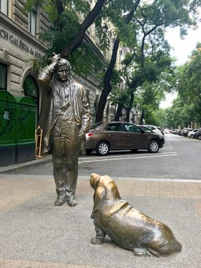 Life-sized bronze statue of Columbo, portrayed in his signature disheveled trench coat, standing and scratching his head in a thoughtful pose. In front of him, there is a smaller statue of his loyal basset hound, sitting and looking up at him. The statues are placed on a sidewalk in an urban area with trees and parked cars lining the street.