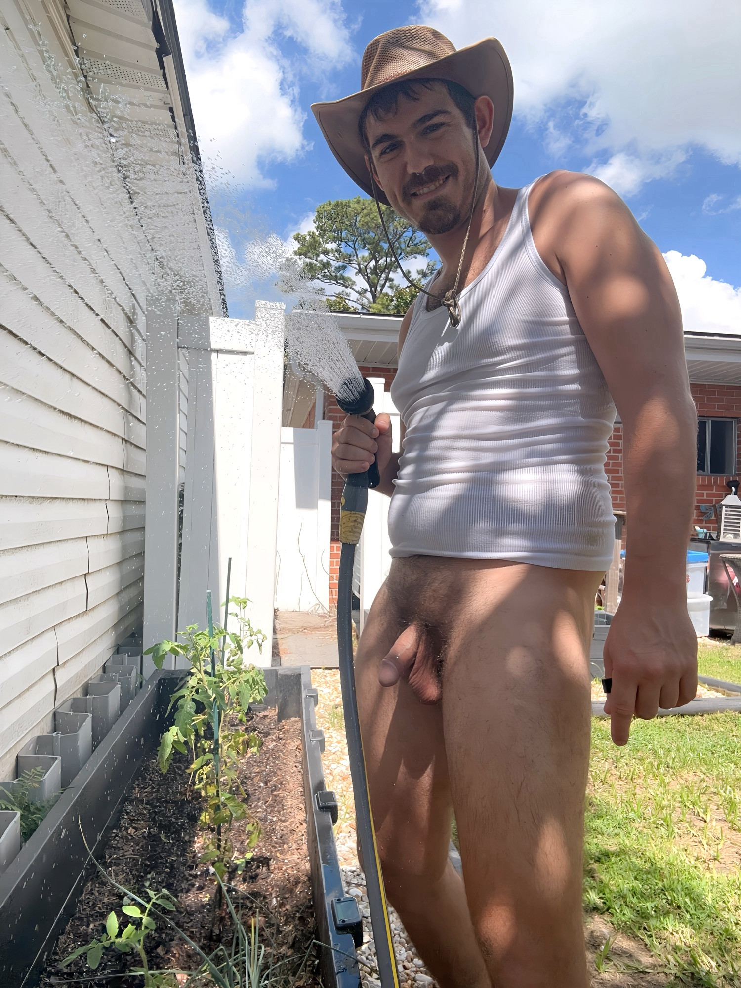 Pup sport with a white tank and bottomless. Hat on. Watering plants outside in the palm tree shade