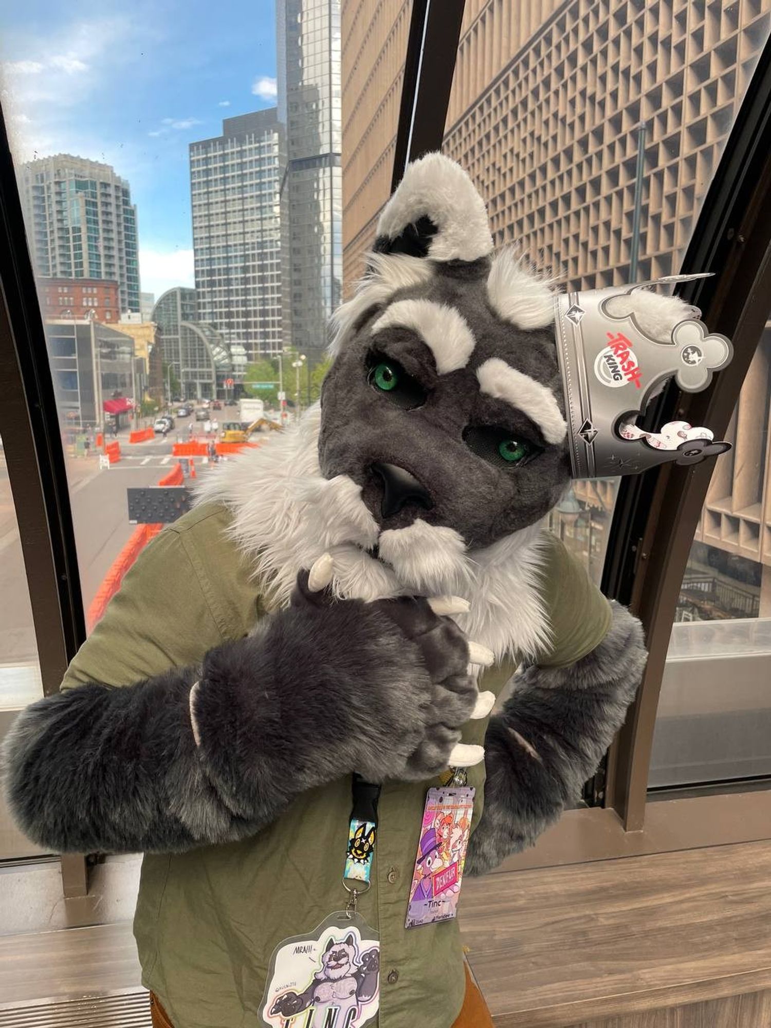 Tinc in a partial fursuit (a grey foxlion head: pointy white tipped ears, green glass eyes, raised white eyebrow, dark Greg fur that goes into a white moustache/beard combo that fades into the mane) wearing a "TrashKing" Burger King style paper crown,  looking at the camera curiously as he tilts his head and strokes his beard