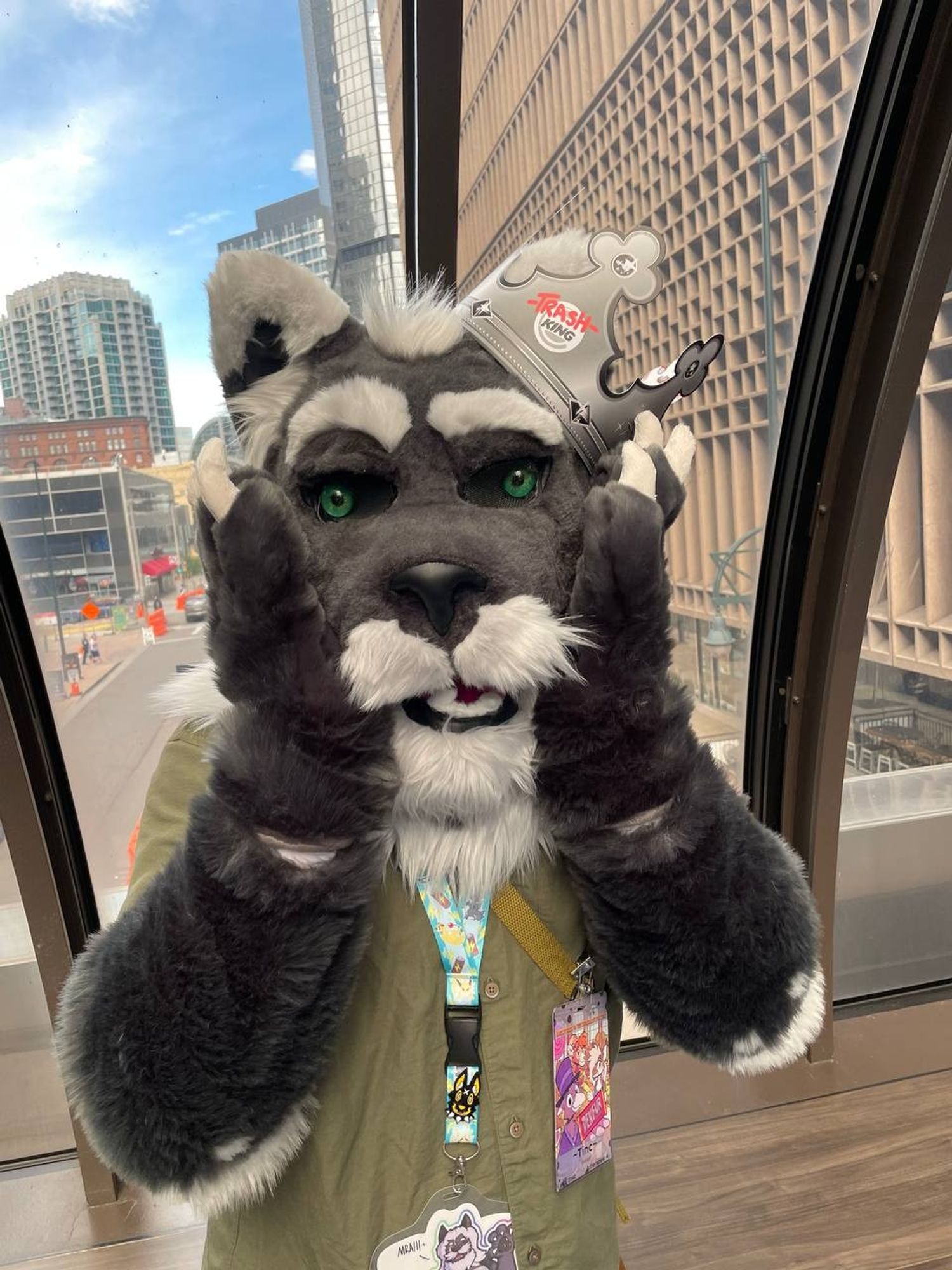 Tinc in a partial fursuit (a grey foxlion head: pointy white tipped ears, green glass eyes, raised white eyebrow, dark Greg fur that goes into a white moustache/beard combo that fades into the mane) wearing a "TrashKing" Burger King style paper crown,  looking at the camera surprised as he slaps his paws to his cheeks