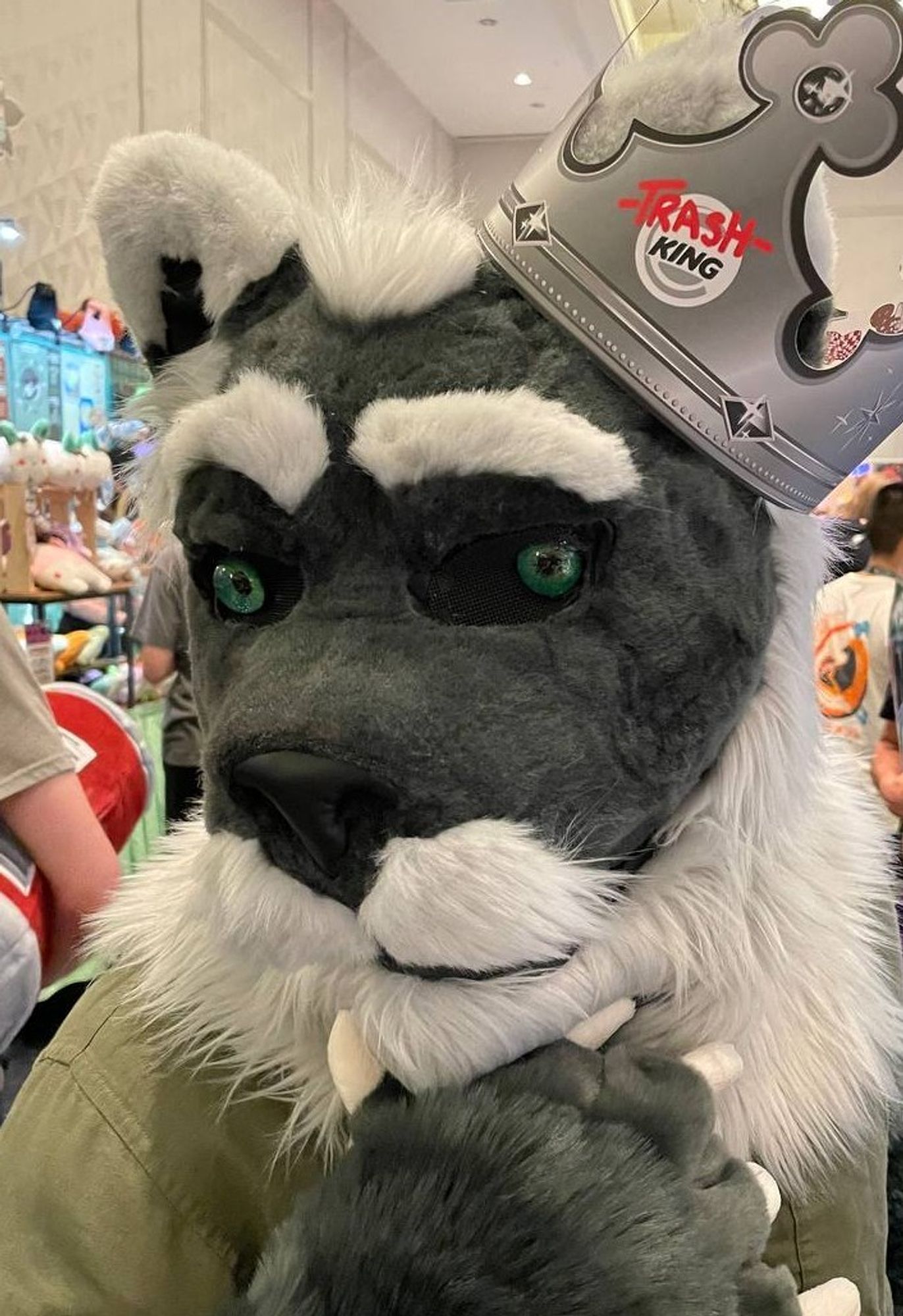 Tinc in a partial fursuit (a grey foxlion head: pointy white tipped ears, green glass eyes, raised white eyebrow, dark Greg fur that goes into a white moustache/beard combo that fades into the mane) wearing a "TrashKing" Burger King style paper crown,  looking pensive past the camera as he strokes his beard. 