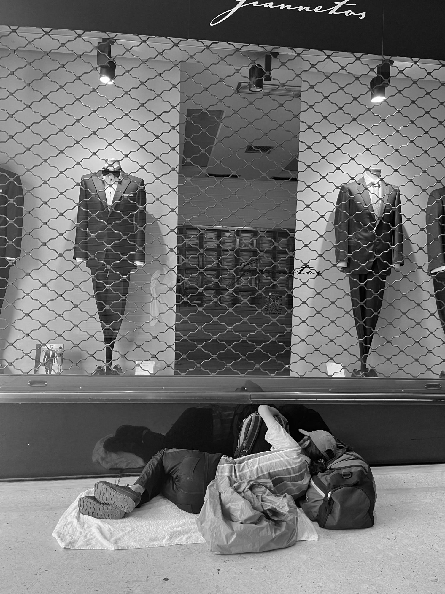 Outside an expensive store displaying high-end men’s suits, two suits spotlight in the centre and seen through the closed security grille. Below, a man is asleep on a towel, clutching his backpack, head pillowed on a duffell , next to his other bagged belongings. He is huddled against the outside of the store, reflected in the black marble frontage, the suits looming over his feet and head.