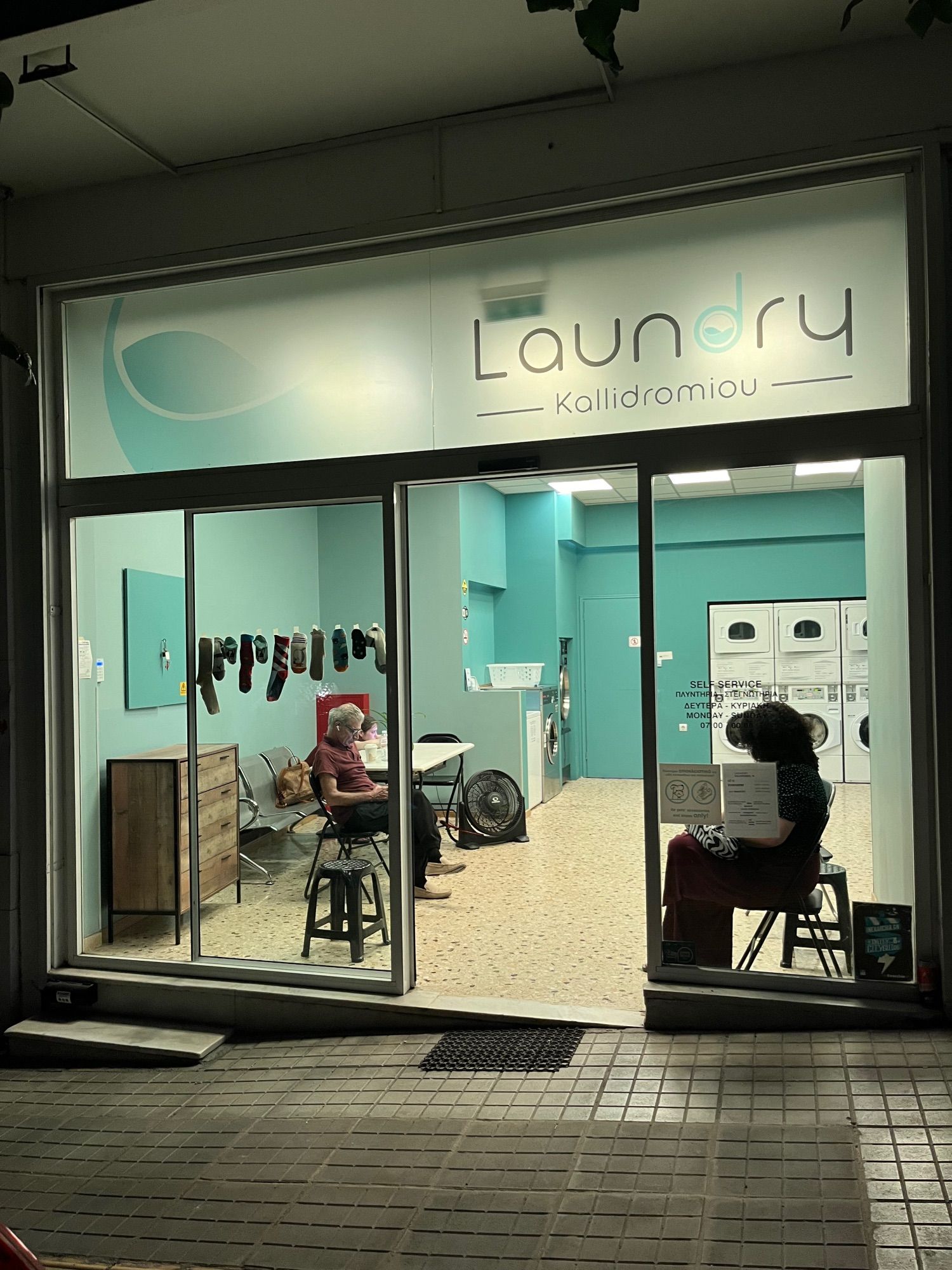 A laundromat with a glass front wall. A wide strip across  the top is frosted and reads Laundry Kalidromiou (a street name) in rounded sans serif letters. Inside the walls are painted pistachio green and the room, brightly lit, glows green into the night. Part of a back of washing machines to the right on the back wall. On the left, someone is slumped across a white topped table. A man in a burgundy t shirt sits on a folding chair, looking at his phone. On the right by the glass wall, another figure slumps on a chair. A row of unmatched socks is taped to the glass.