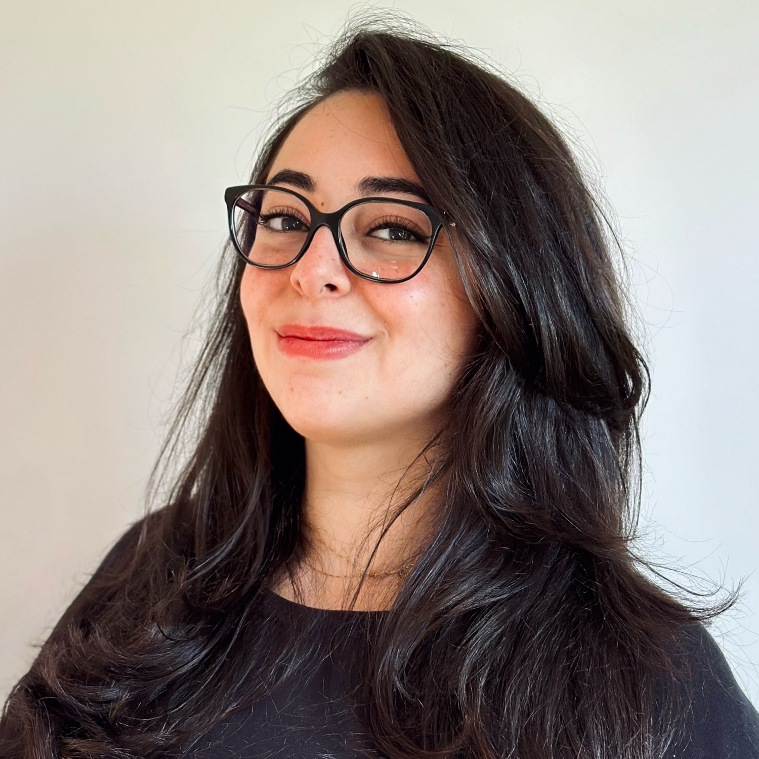 Photograph of a woman with light olive skin. She has long dark hair, brown eyes and black framed glasses. She is smiling while looking at the camera.