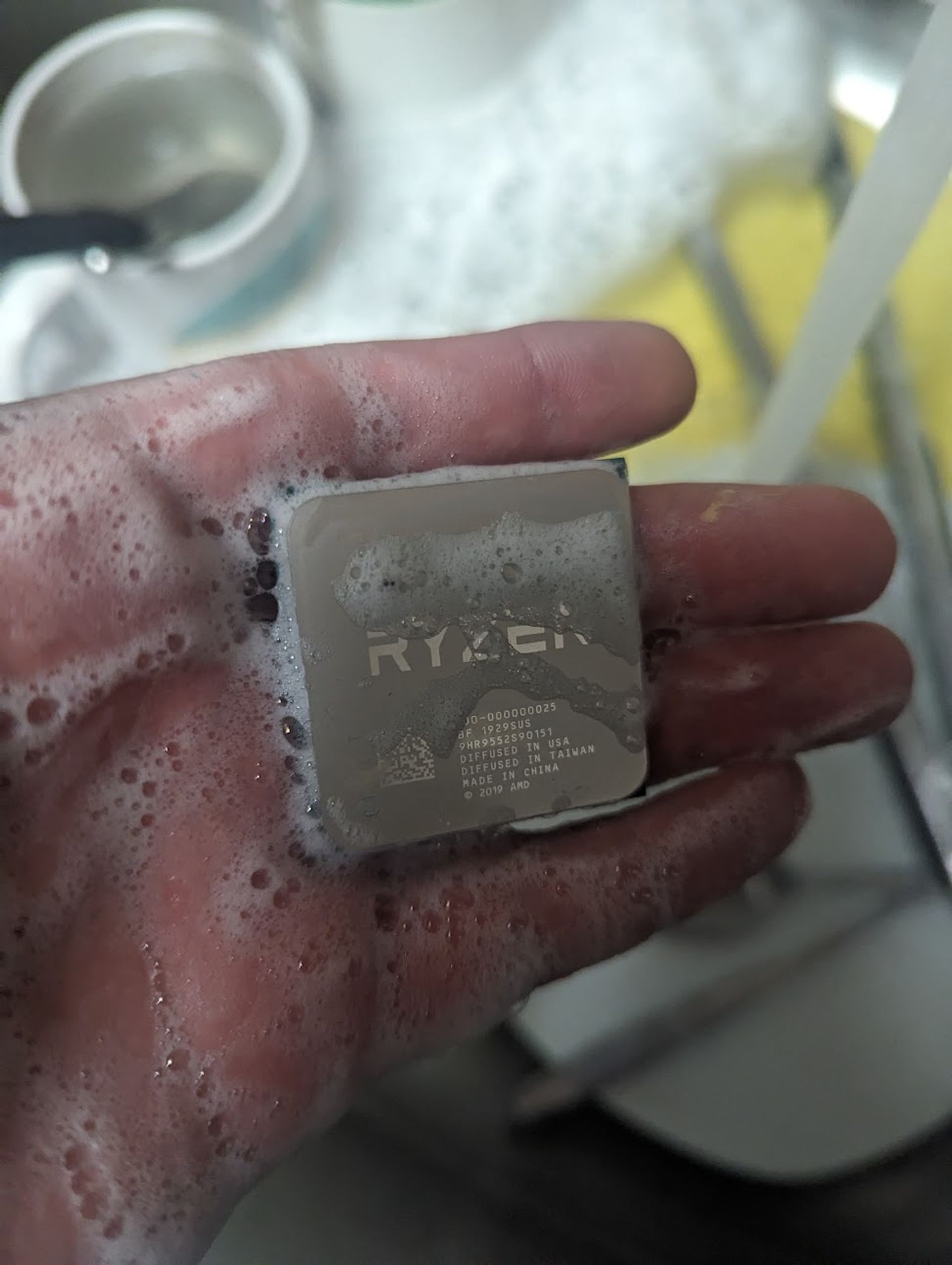 an AMD ryzen CPU being held above a sink, the hand and cpu are both wet and covered in soap suds, there's a blurry sink with some dishes and a running tap in the background.