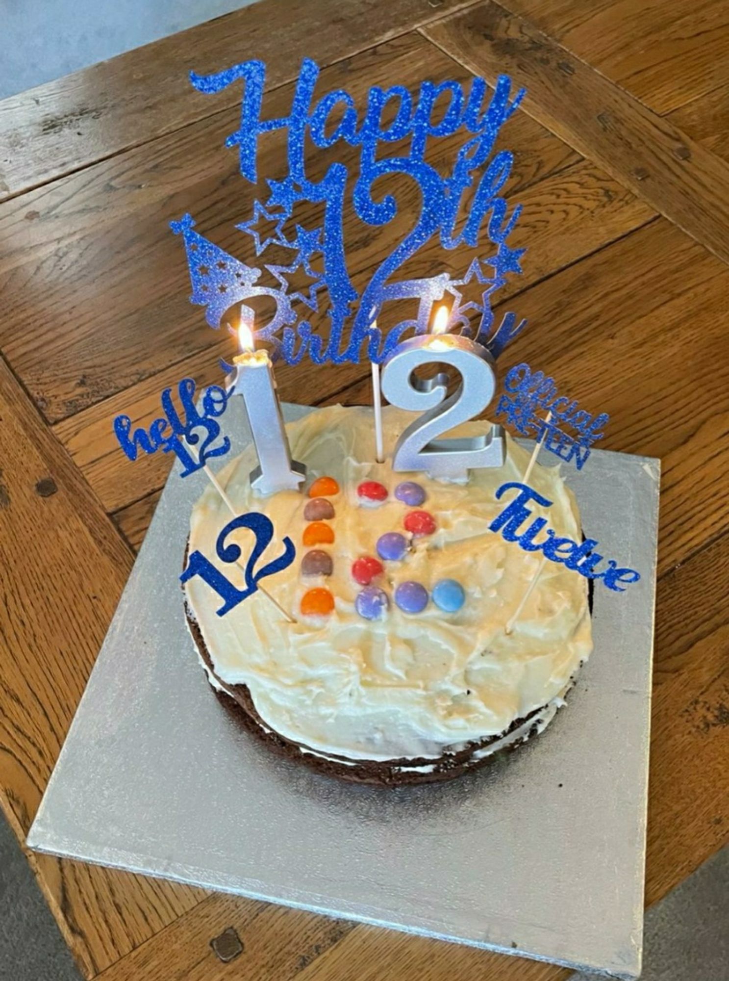 Chocolate birthday cake with cream frosting. The number 12 is marked out in Smarties. There are two silver candles, a 1 and a 2. At the back a blue decoration saying Happy 12th birthday