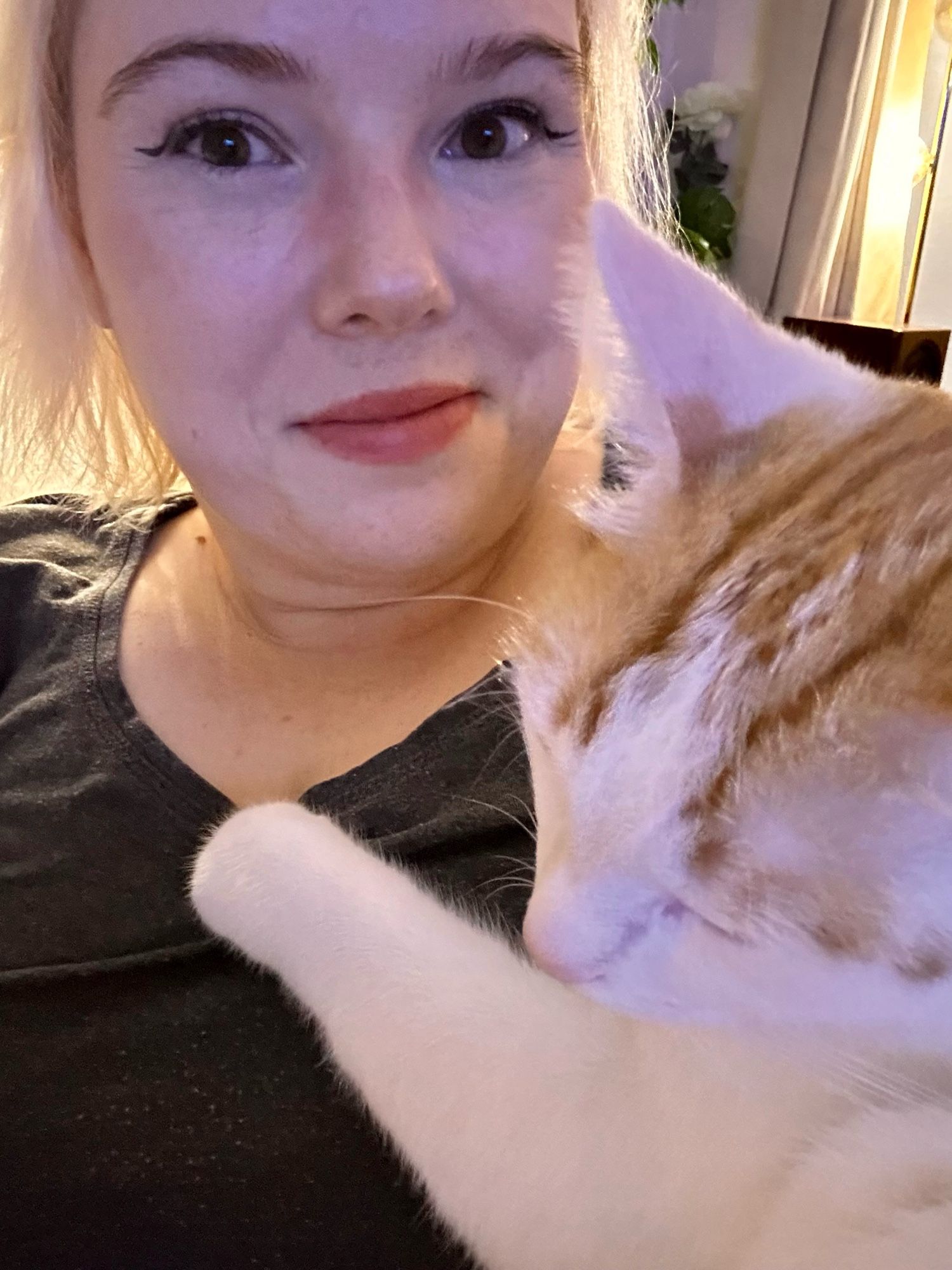 A photo of me, a white woman with shoulder length blonde hair, smiling at the camera, while a white and orange cat is snoozing on my chest