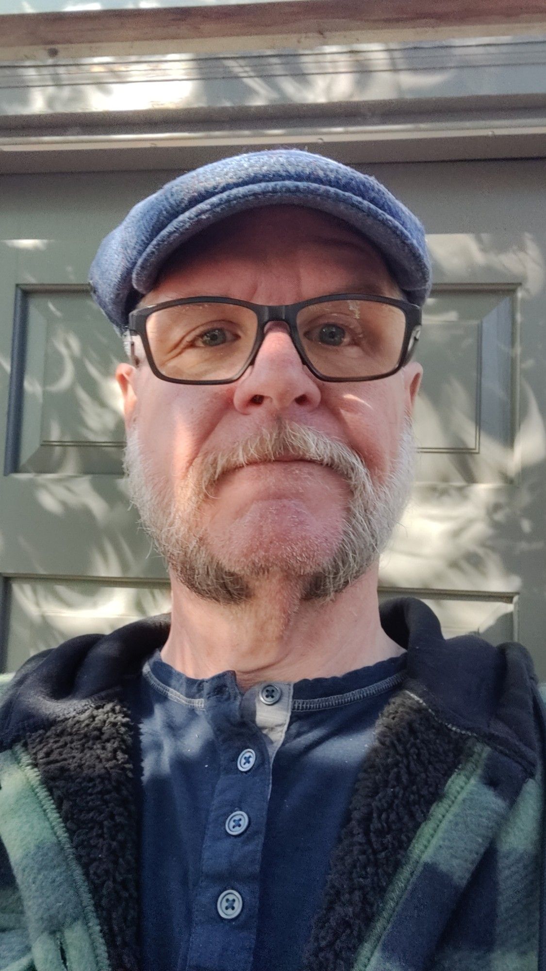 Selfie. Wearing plaid microfiber hoodie overshirt (forest green and black) and a blue flat cap. Hair is a medium fade cut. Facial hair is trimmed to mutton chops connected by mustache. The shadows in the background have crescent shapes due to the eclipse.