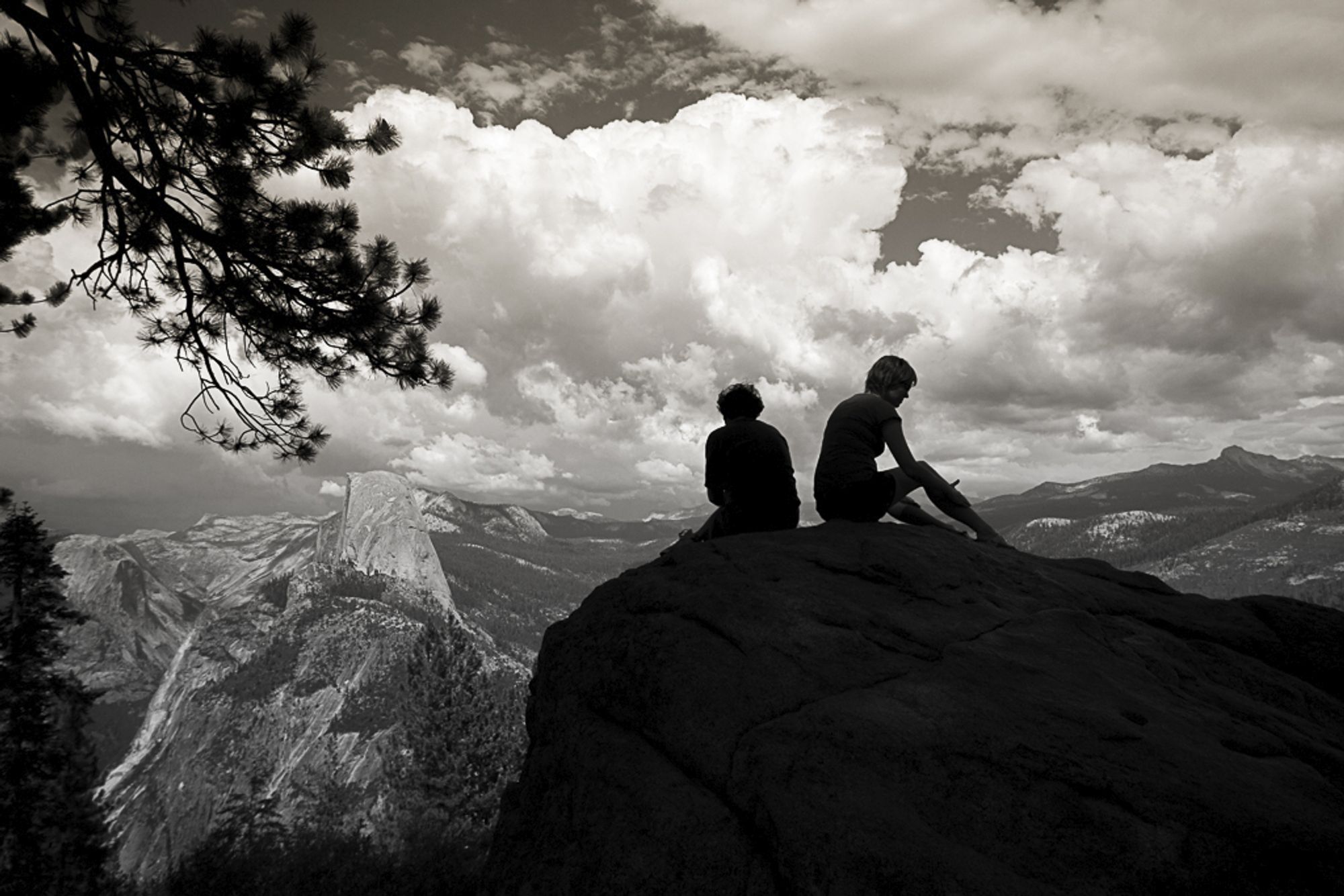 In the foreground are two people sitting on a rock silhouetted against a sky with threatening thunderheads. In the distance on the left is Half Dome, Clouds Rest, and the high Sierra. A pine branch reaches down from the left as if to hug Half Dome.