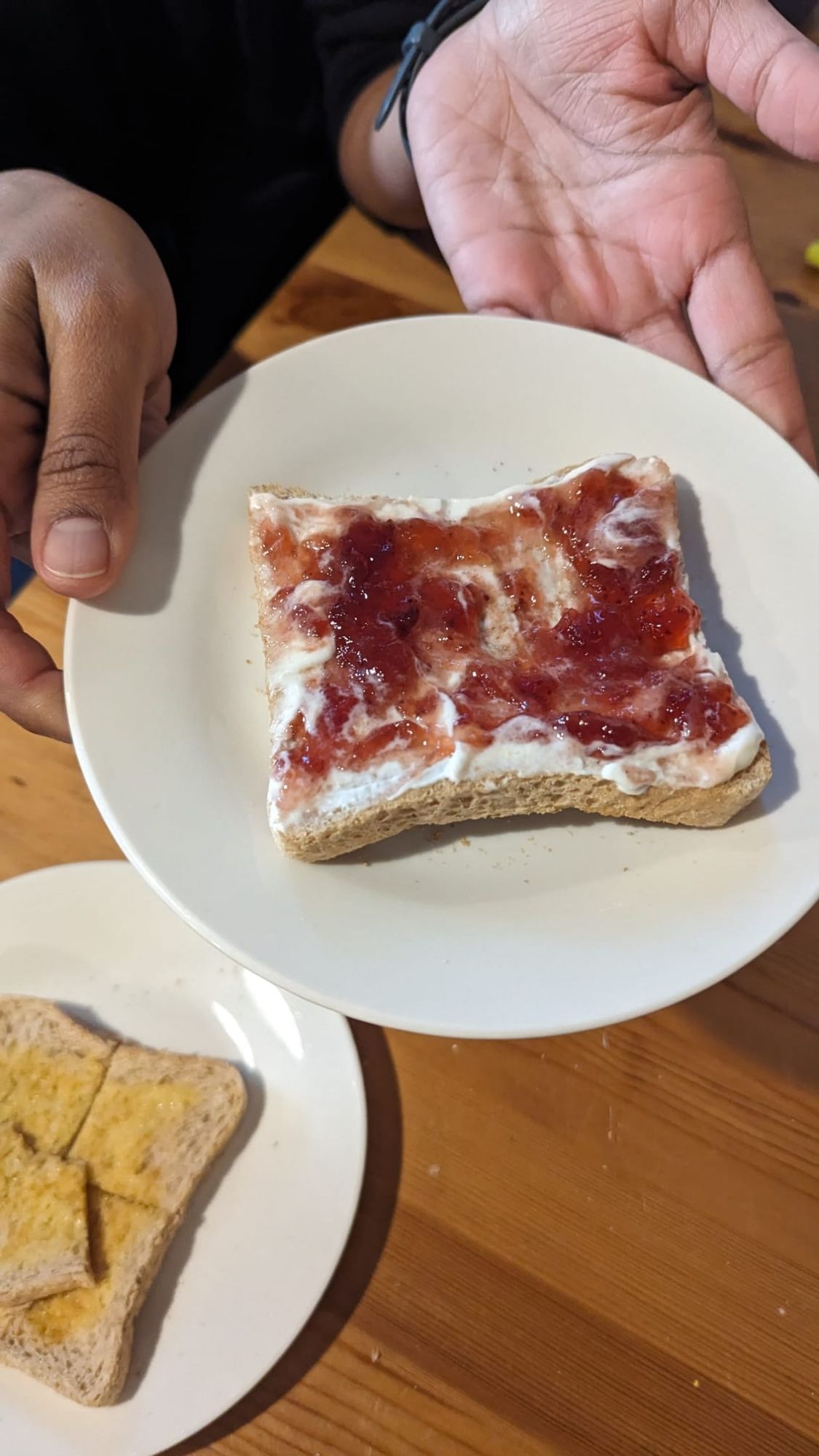 A picture of someone holding a white plate with toast on it. The toast has a lashing of fromage frais and strawberry jam on it.
