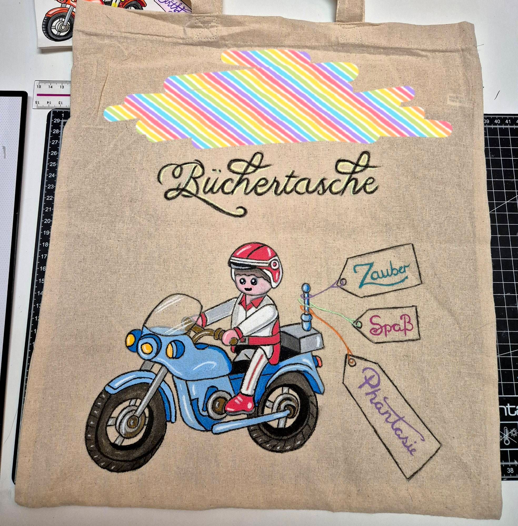 Eine beige Stofftasche. Gemaltes Motiv ist ein blaues Playmobilmotorrad, auf dem eine Playmobilfigur mit weiß/roter Motorradkluft sitzt. An der Antenne hängen 3 Anhänger mit den Worten "Zauber", "Spaß" und Phantasie.
Über dem Bild steht in geschwungener Calligraphie Schrift "Büchertasche", darüber wurde der Name des Kindes bunt verpixelt.